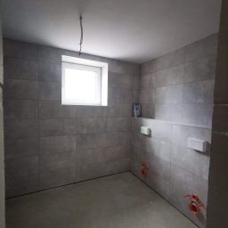 Arton - Surowa łazienka w trakcie wykańczania: szare płytki na ścianach, wnęka z białymi elementami podtynkowymi, wystające rury instalacji hydraulicznej, surowy beton na podłodze, pojedynczy kabel...