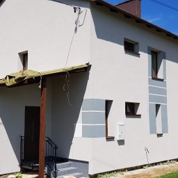 Arton - Dwukondygnacyjny dom z jasną elewacją i szarymi akcentami wokół okien, częściowo osłonięty zieloną plandeką nad wejściem, widok z zewnątrz w słoneczny dzień.