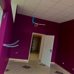 Arton - Wnętrze pokoju po malowaniu ścian na intensywny fiolet, z widocznymi instalacjami elektrycznymi i wentylacyjnymi przed zabudową, oraz częściowo ułożoną podłogą z płytek w odcieniach beżu i brązu.