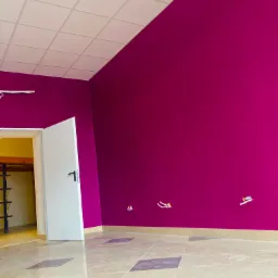 Świeżo pomalowane wnętrze z fuksjową ścianą, widoczne wystające kable elektryczne, kremowa podłoga z ciemnymi wstawkami i biały sufit kasetonowy.