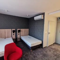 Pokój hotelowy z dwoma pojedynczymi łóżkami, czerwonym fotelem, wzorzystą tapetą i klimatyzacją, oświetlony światłem dziennym wpadającym przez okno.