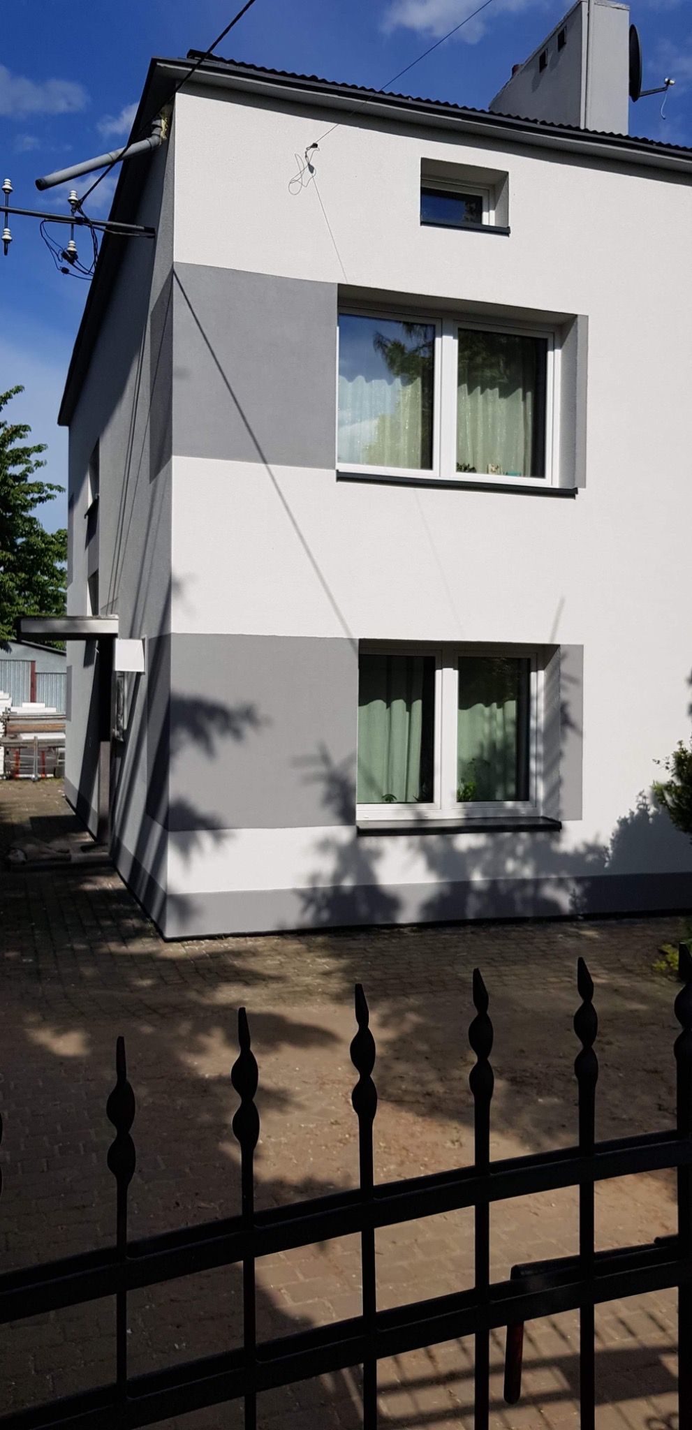 Dwukondygnacyjny dom z nową, dwukolorową elewacją w odcieniach szarości i bieli, widoczny zza ozdobnego, metalowego ogrodzenia.