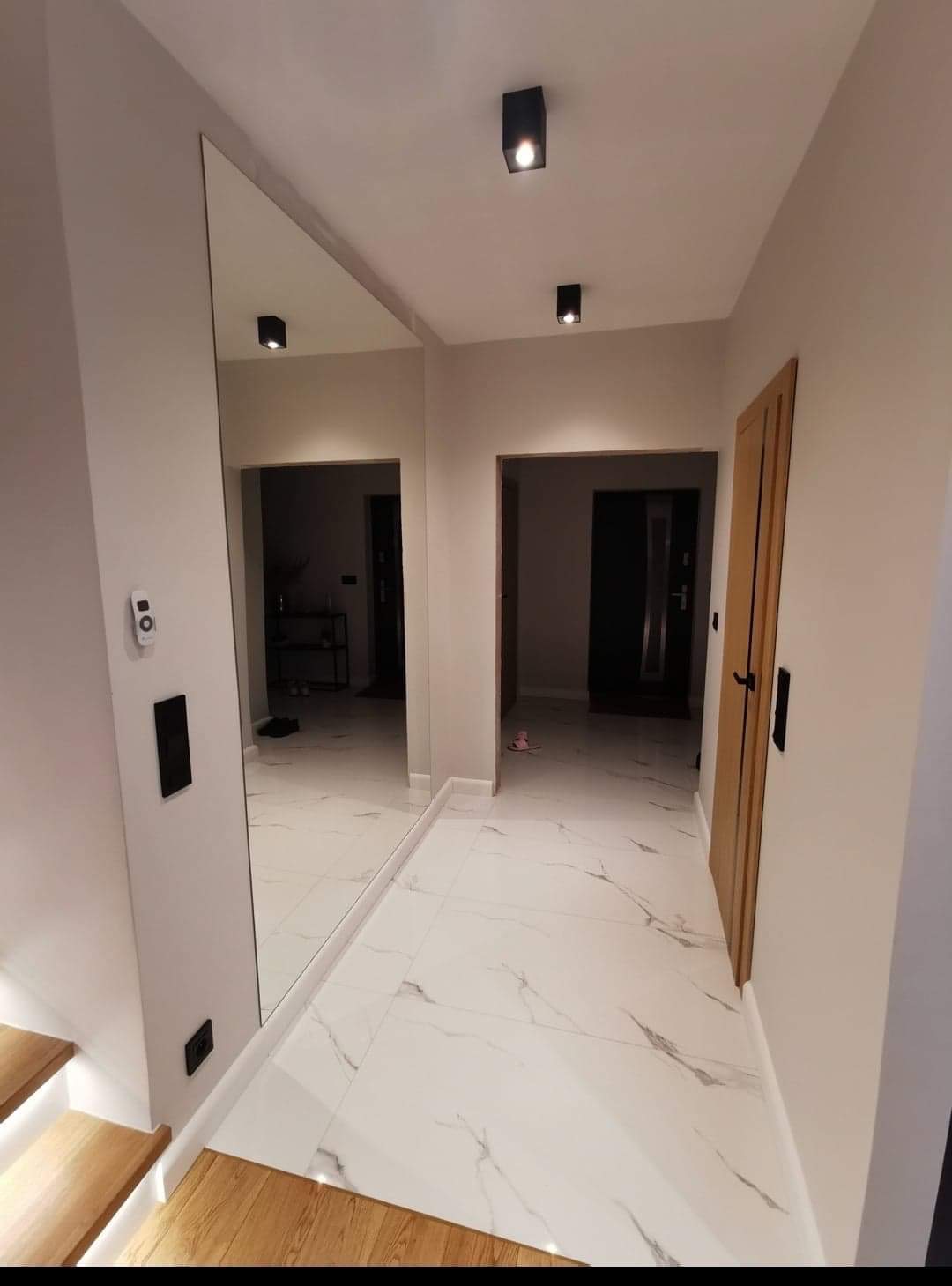 Nowoczesny przedpokój z białymi marmurowymi płytkami podłogowymi, dużym lustrem na ścianie i drewnianymi schodami z podświetleniem LED.