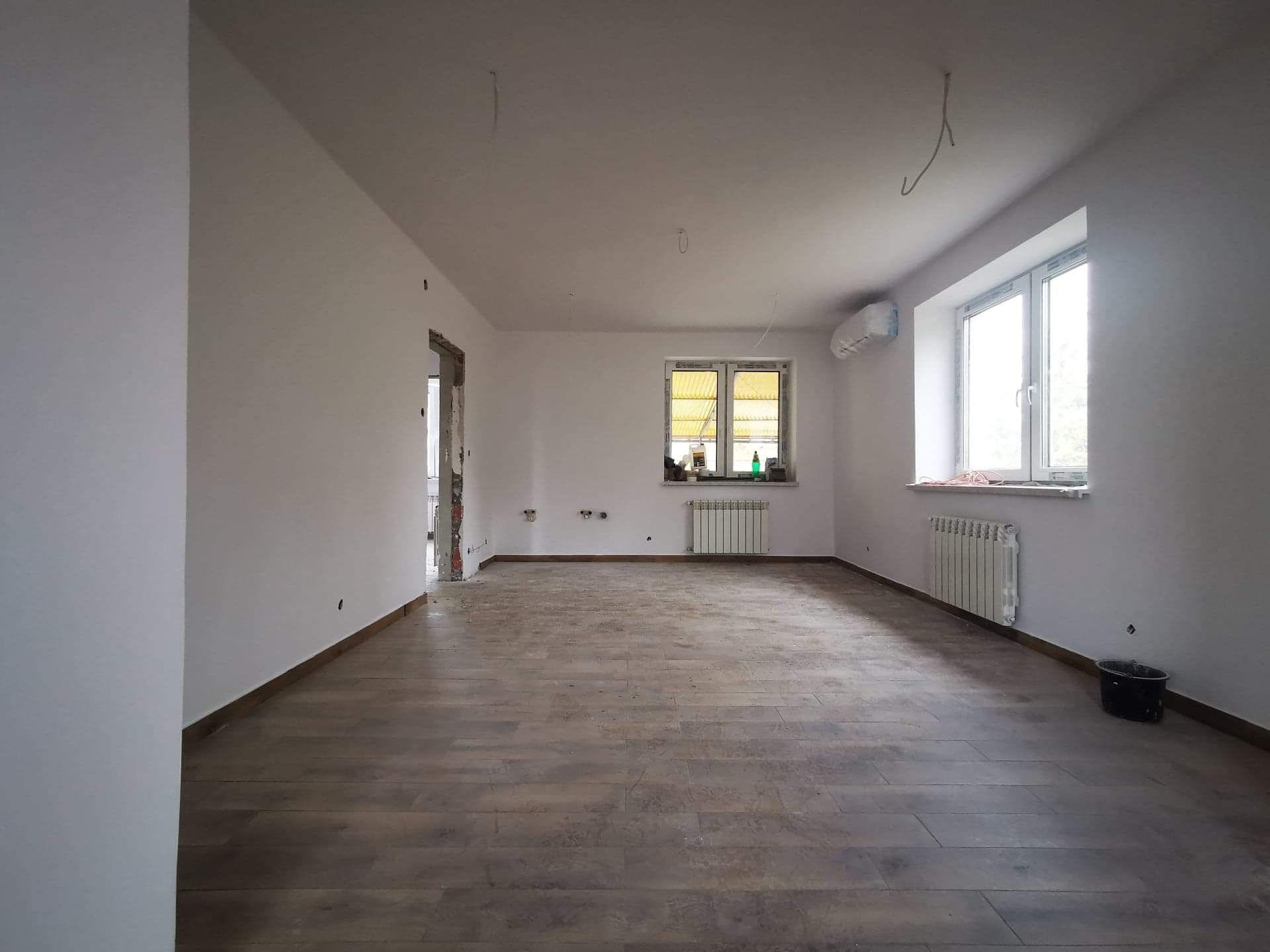 Wnętrze pokoju po położeniu drewnopodobnych płytek podłogowych, świeżo pomalowane ściany na biało, grzejniki pod oknami, widoczne ślady prac wykończeniowych.