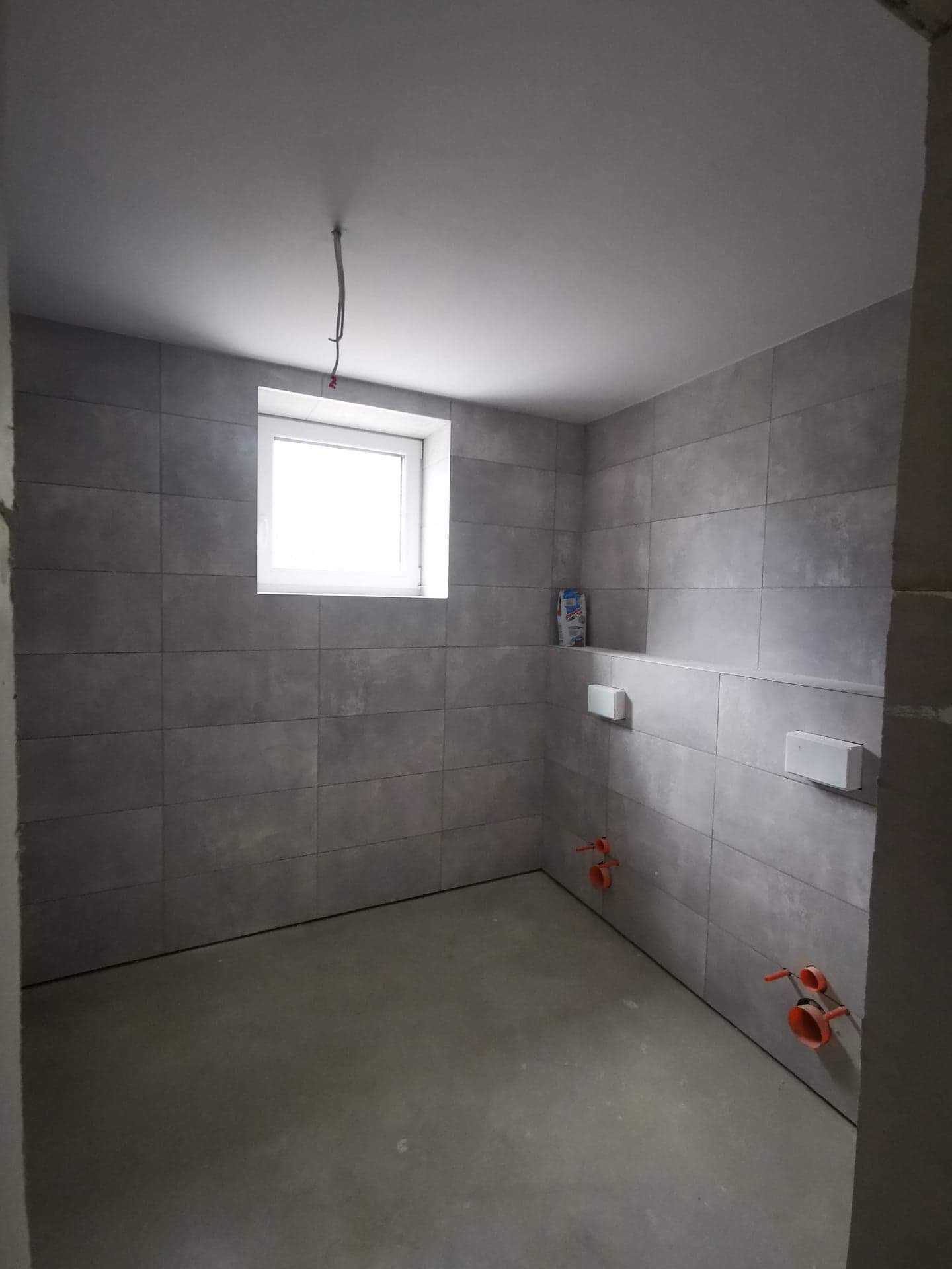 Surowa łazienka w trakcie wykańczania: szare płytki na ścianach, wnęka z białymi elementami podtynkowymi, wystające rury instalacji hydraulicznej, surowy beton na podłodze, pojedynczy kabel...