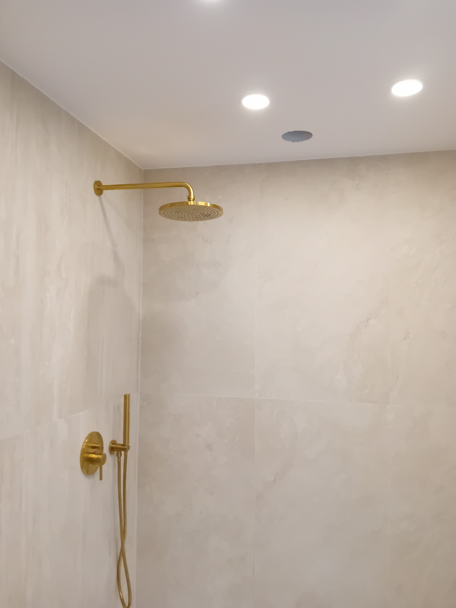 Wykończona łazienka z beżowymi płytkami imitującymi marmur i złotą armaturą: deszczownica i bateria podtynkowa. W suficie zamontowane trzy halogenowe lampy.