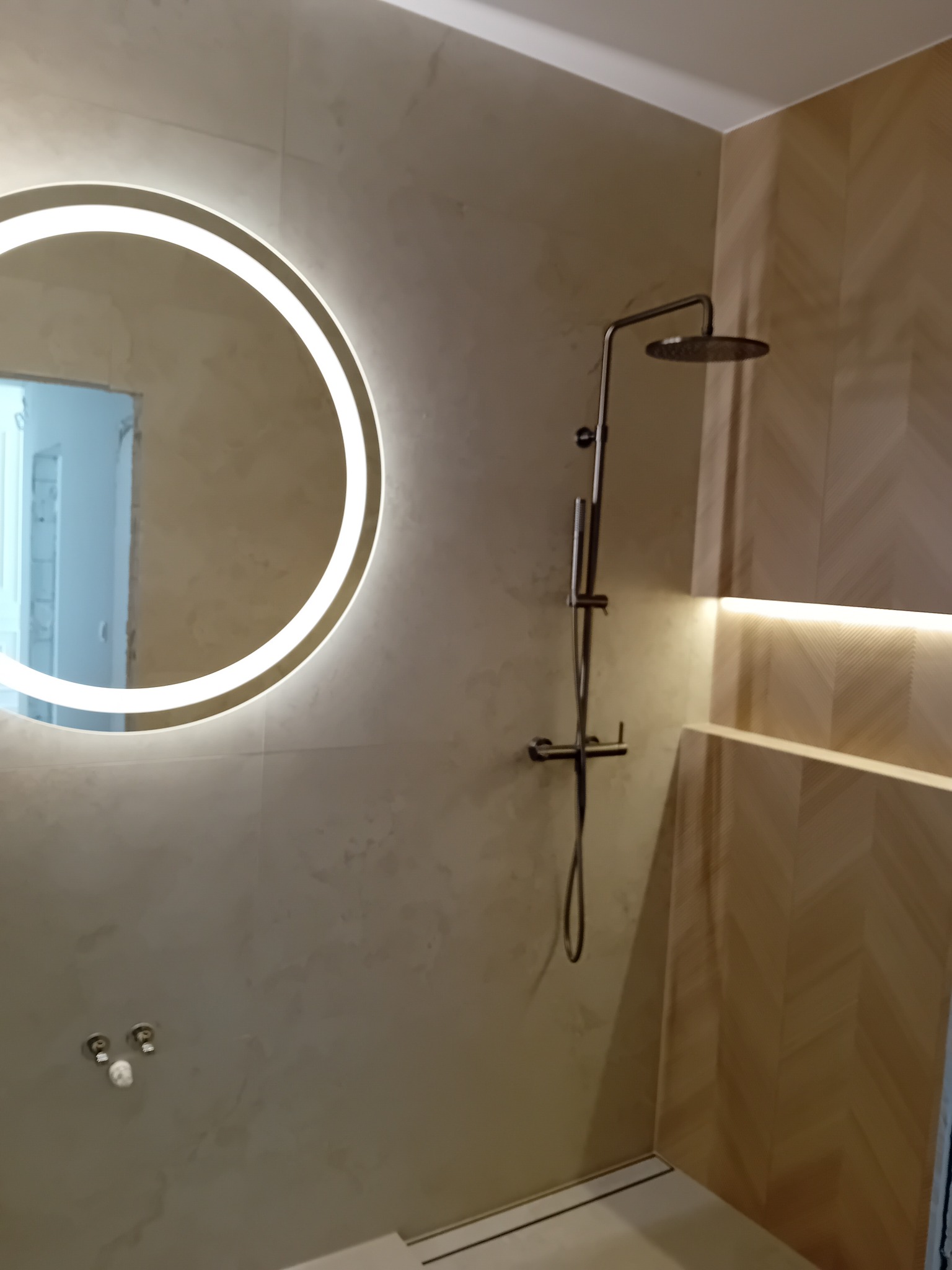 Nowoczesna łazienka z okrągłym lustrem podświetlanym LED, deszczownicą i płytkami imitującymi drewno ułożonymi w jodełkę.