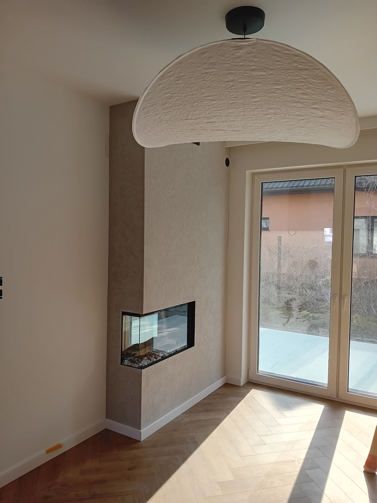 Nowoczesny salon z narożnym kominkiem obudowanym szarym kamieniem dekoracyjnym, drewnianą podłogą ułożoną w jodełkę i dużym oknem balkonowym.