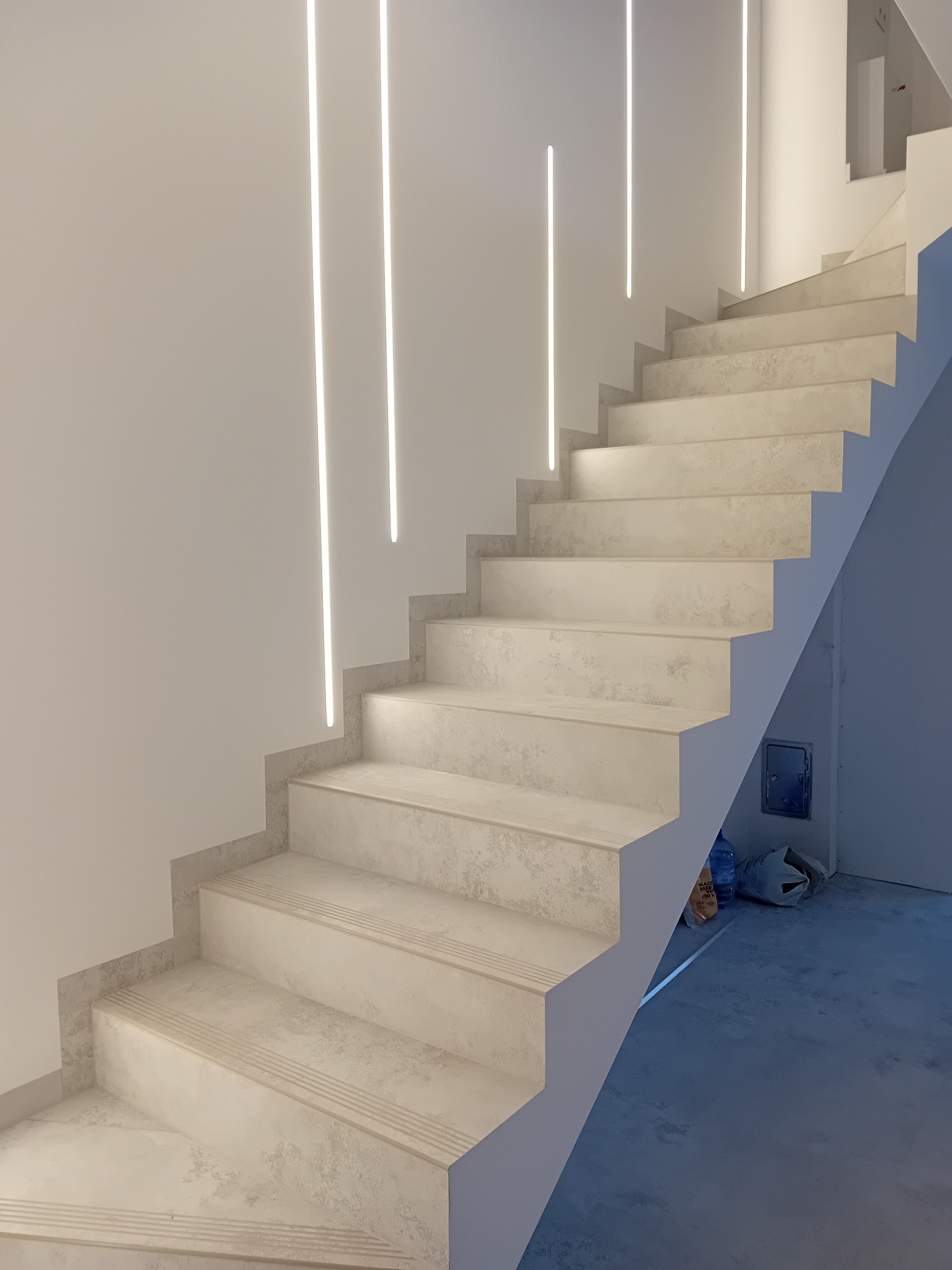 Nowoczesne schody z podświetleniem LED w ścianie, wykończone płytkami imitującymi kamień, z widocznymi torbami i butelką na podłodze.