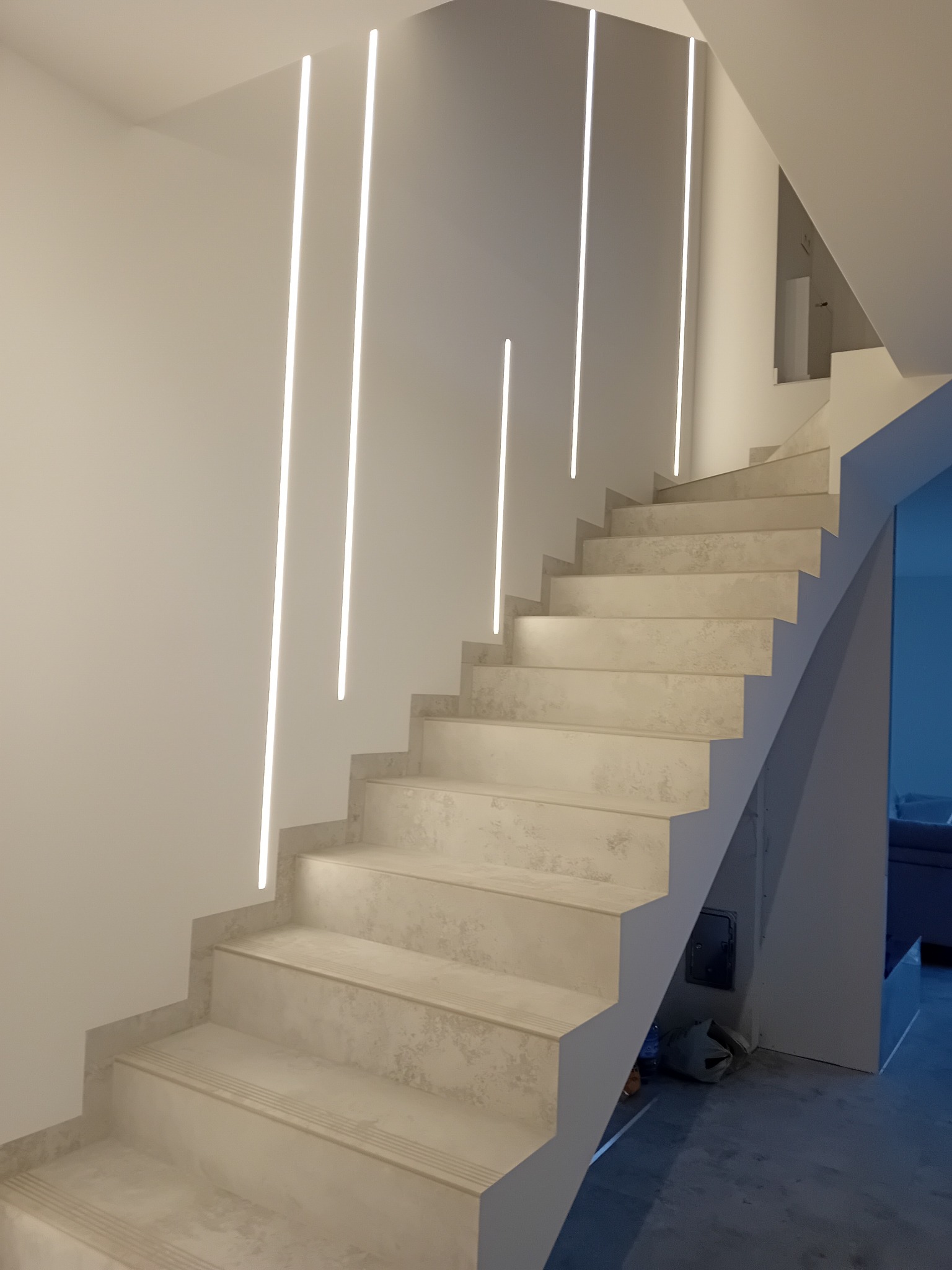 Nowoczesne schody z podświetleniem LED wzdłuż ściany, wykończone w jasnych odcieniach szarości, z widocznymi śladami prac wykończeniowych na podłodze.