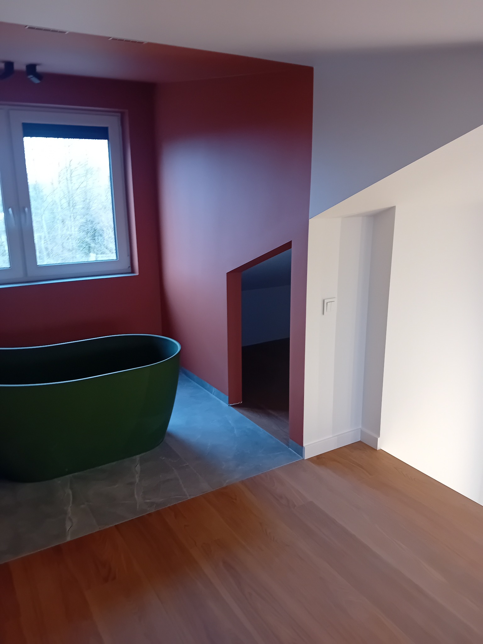 Nowoczesna łazienka z wolnostojącą, oliwkową wanną, ściany pomalowane na kolor terakoty, podłoga wyłożona płytkami imitującymi marmur oraz drewnianym parkietem.