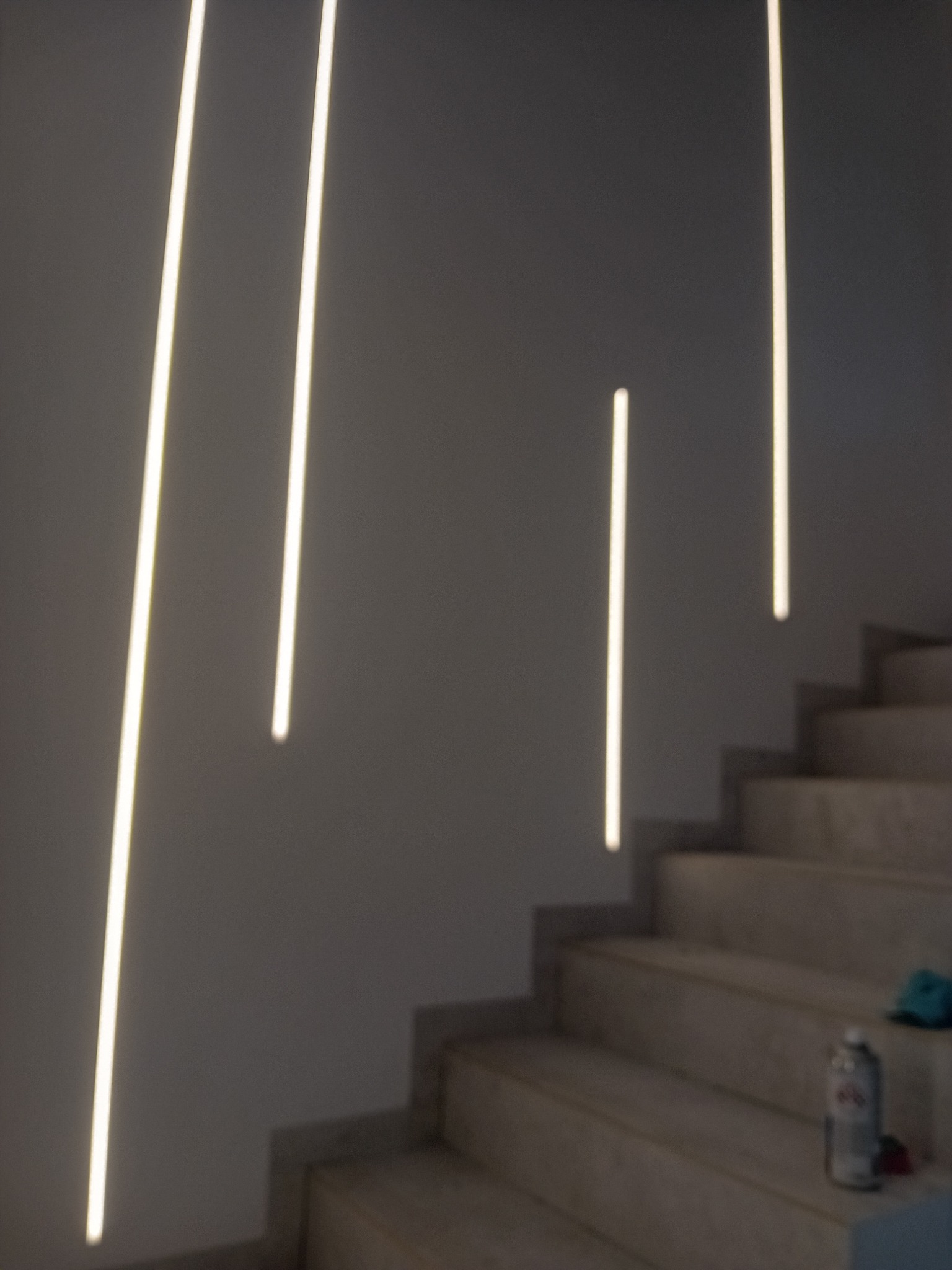 Minimalistyczne oświetlenie schodów z pionowymi liniami LED wpuszczonymi w ścianę, widoczny fragment schodów z butelką i szmatką.