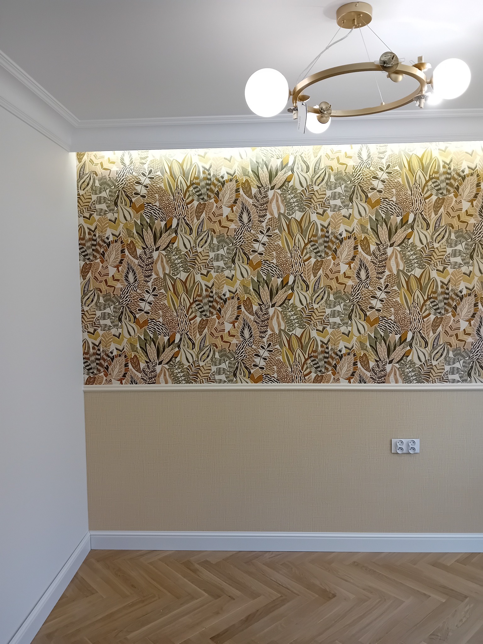 Pokój po remoncie z wzorzystą tapetą w odcieniach brązu i zieleni, kremową dolną częścią ściany, parkietem w jodełkę i nowoczesną, złotą lampą sufitową z białymi kulami.