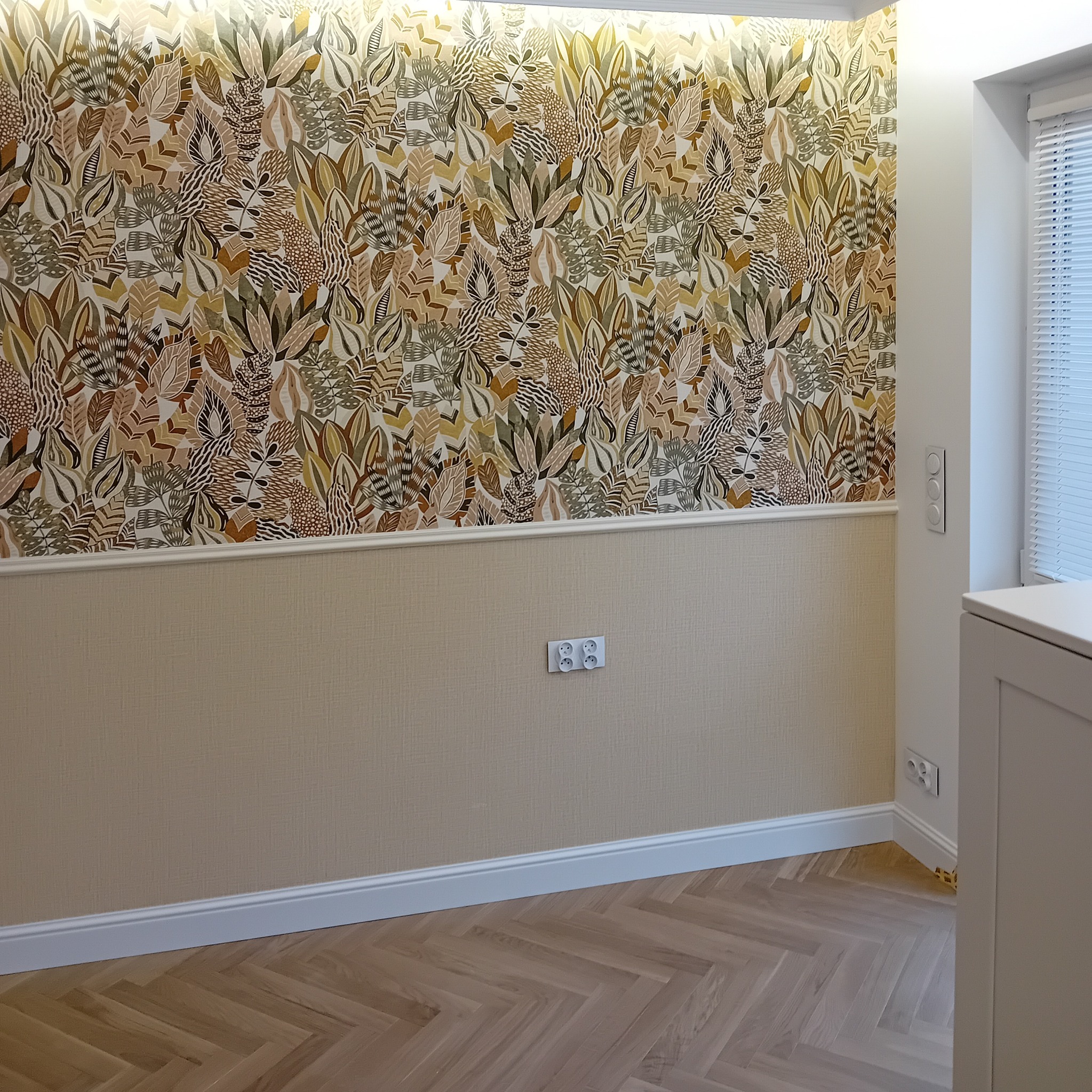 Pokój po remoncie z dekoracyjną tapetą w liście, lamperią w odcieniach beżu, białymi listwami przypodłogowymi i parkietem w jodełkę