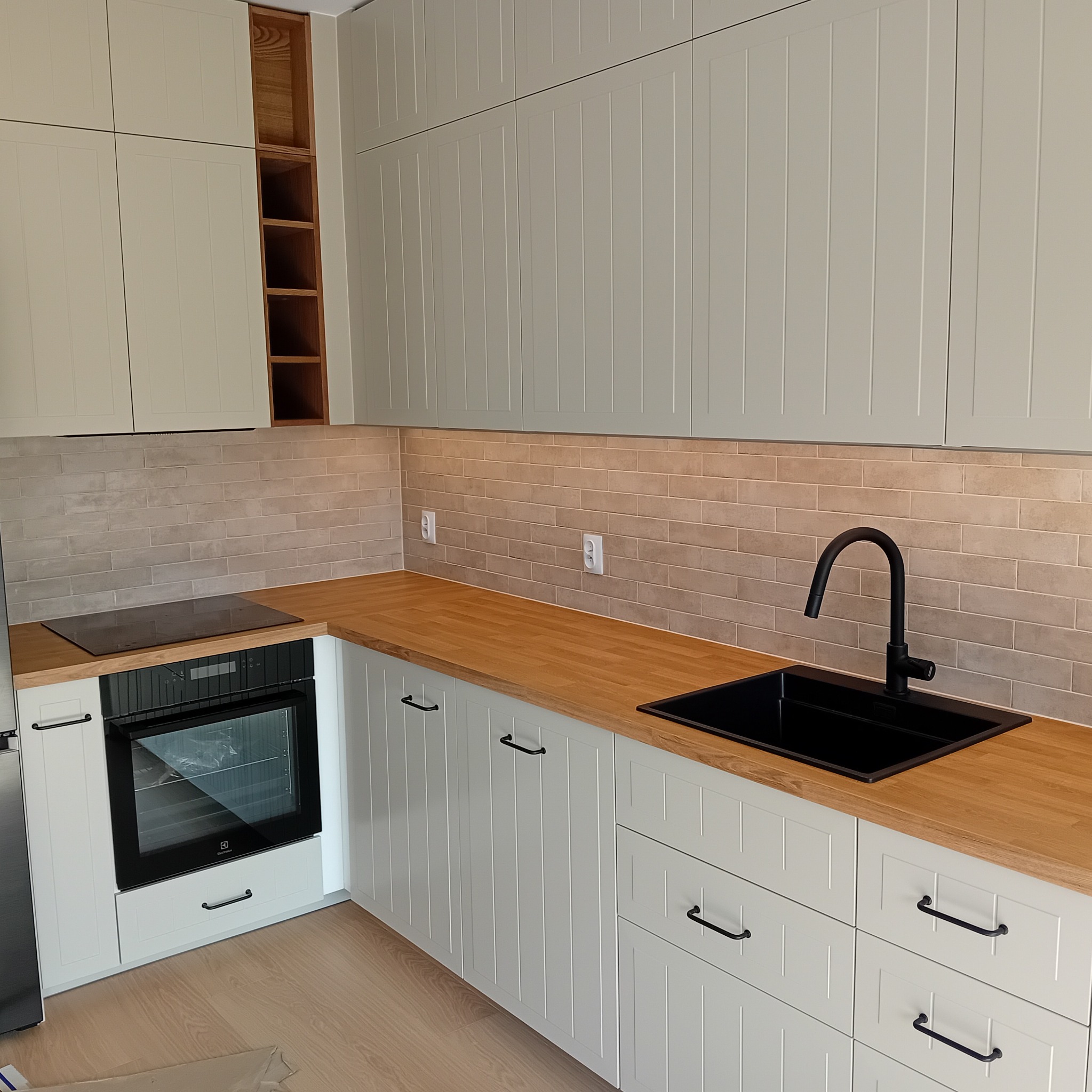 Jasna kuchnia w stylu rustykalnym z drewnianym blatem, czarnym zlewem i armaturą, oraz płytkami imitującymi cegłę na ścianie.