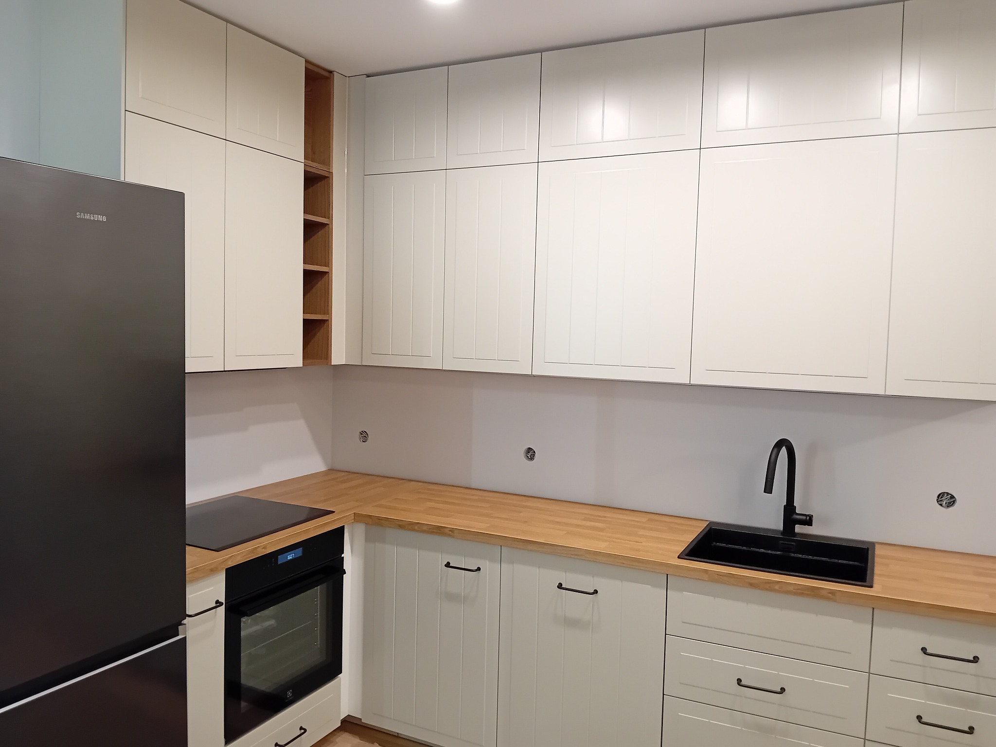 Nowoczesna kuchnia z białymi szafkami z frezowanymi frontami, drewnianym blatem, czarnym zlewem i czarną lodówką Samsung.