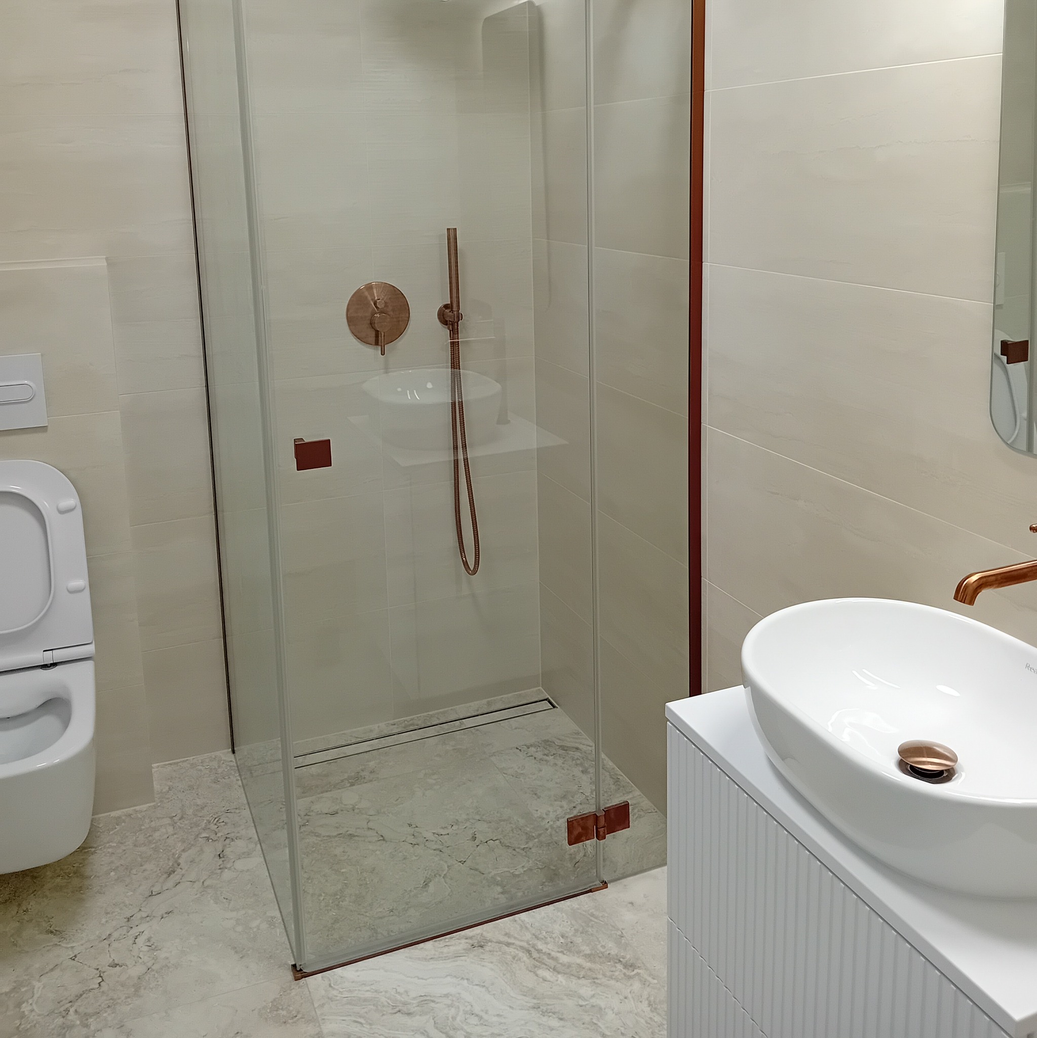 Nowoczesna łazienka z kabiną prysznicową typu walk-in, umywalką nablatową na białej szafce z ryflowanym frontem i armaturą w kolorze miedzi.