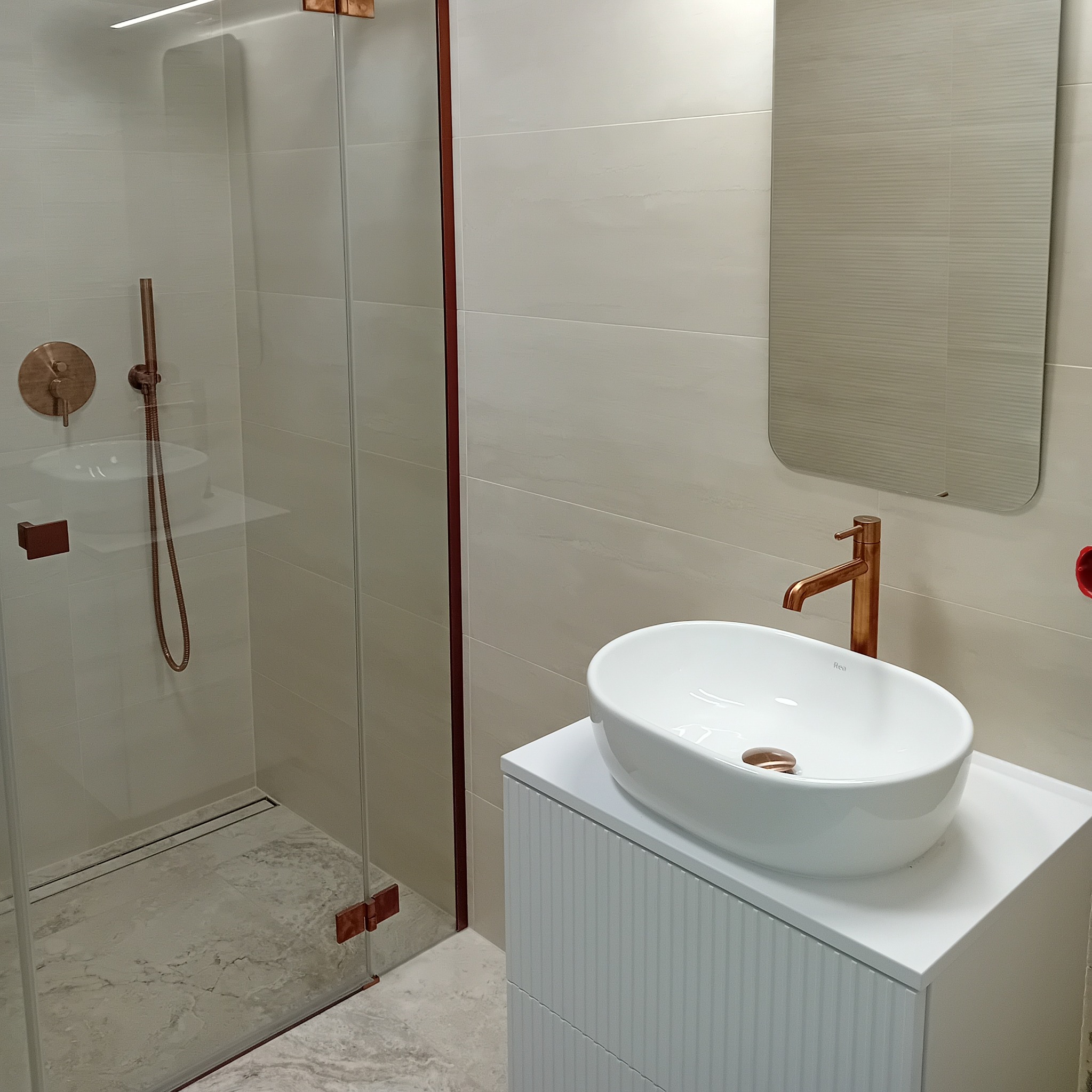 Nowoczesna łazienka z kabiną prysznicową, umywalką nablatową na białej szafce z frezowanymi frontami i armaturą w kolorze miedzi.