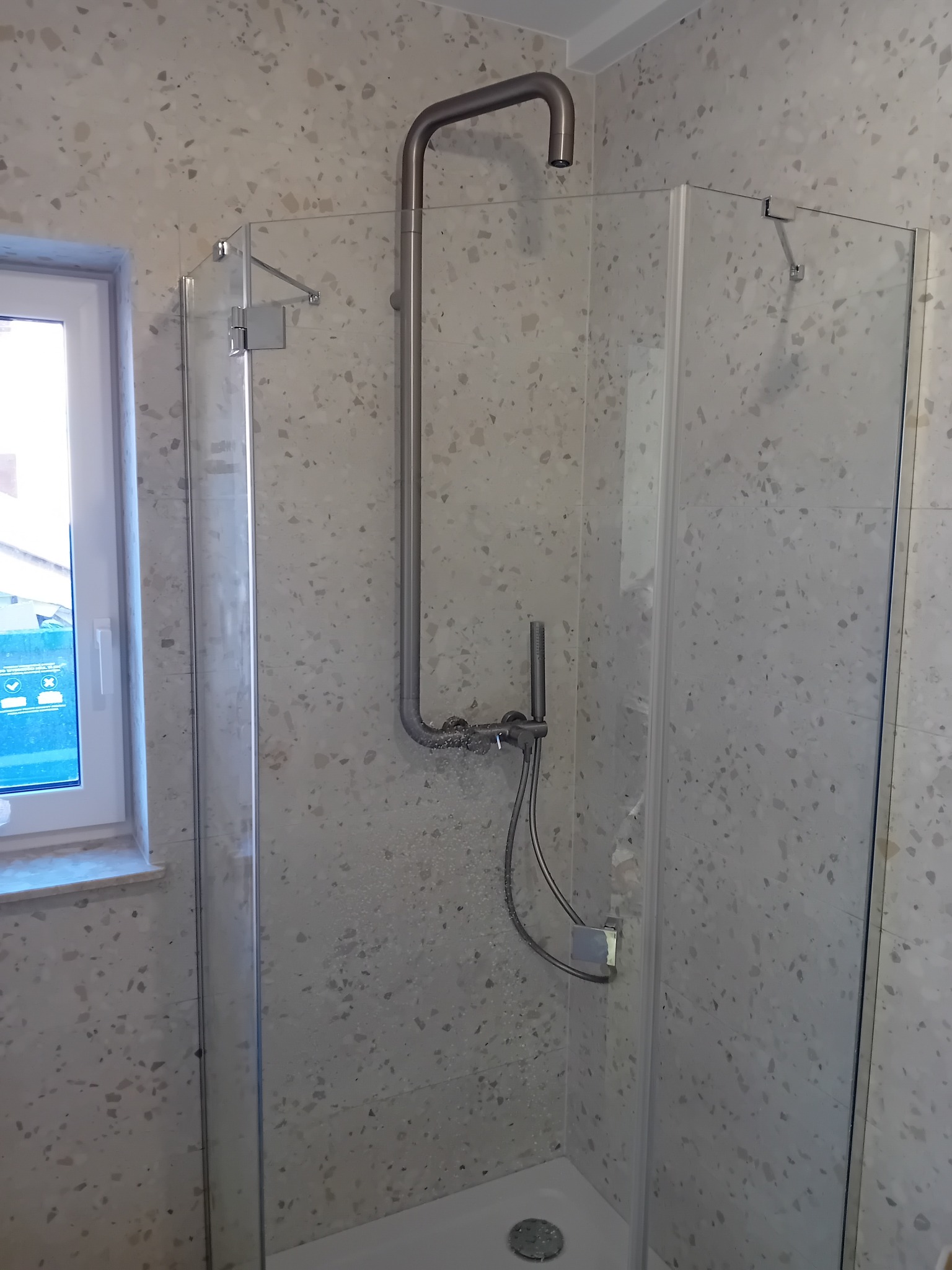 Nowoczesna kabina prysznicowa z szklanymi ścianami i deszczownicą, wykończona płytkami imitującymi lastryko, widok z boku na fragment okna.