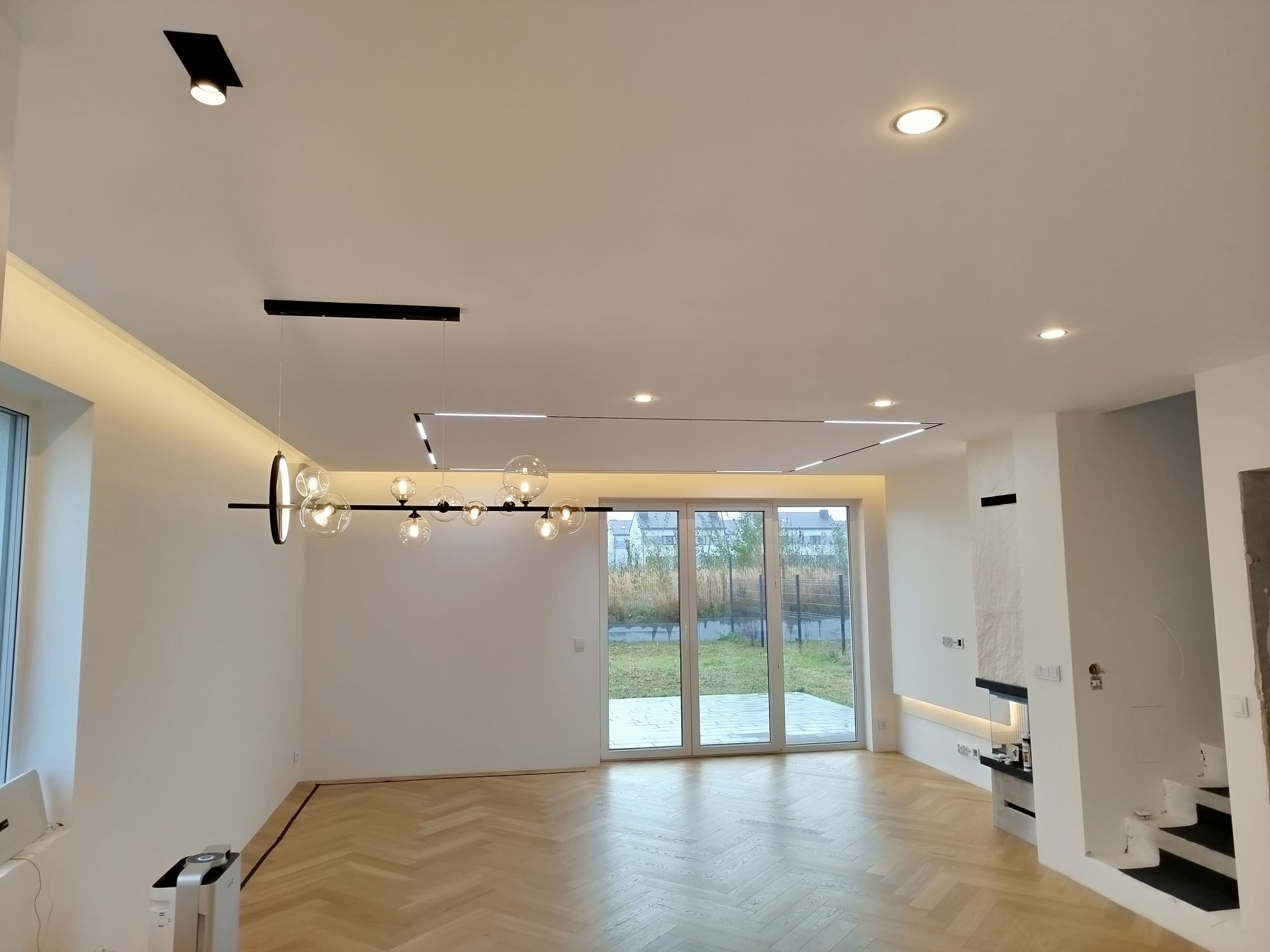 Jasne, przestronne wnętrze po remoncie: parkiet w jodełkę, białe ściany, nowoczesne oświetlenie sufitowe i kominek. Widok na ogród przez duże okna.