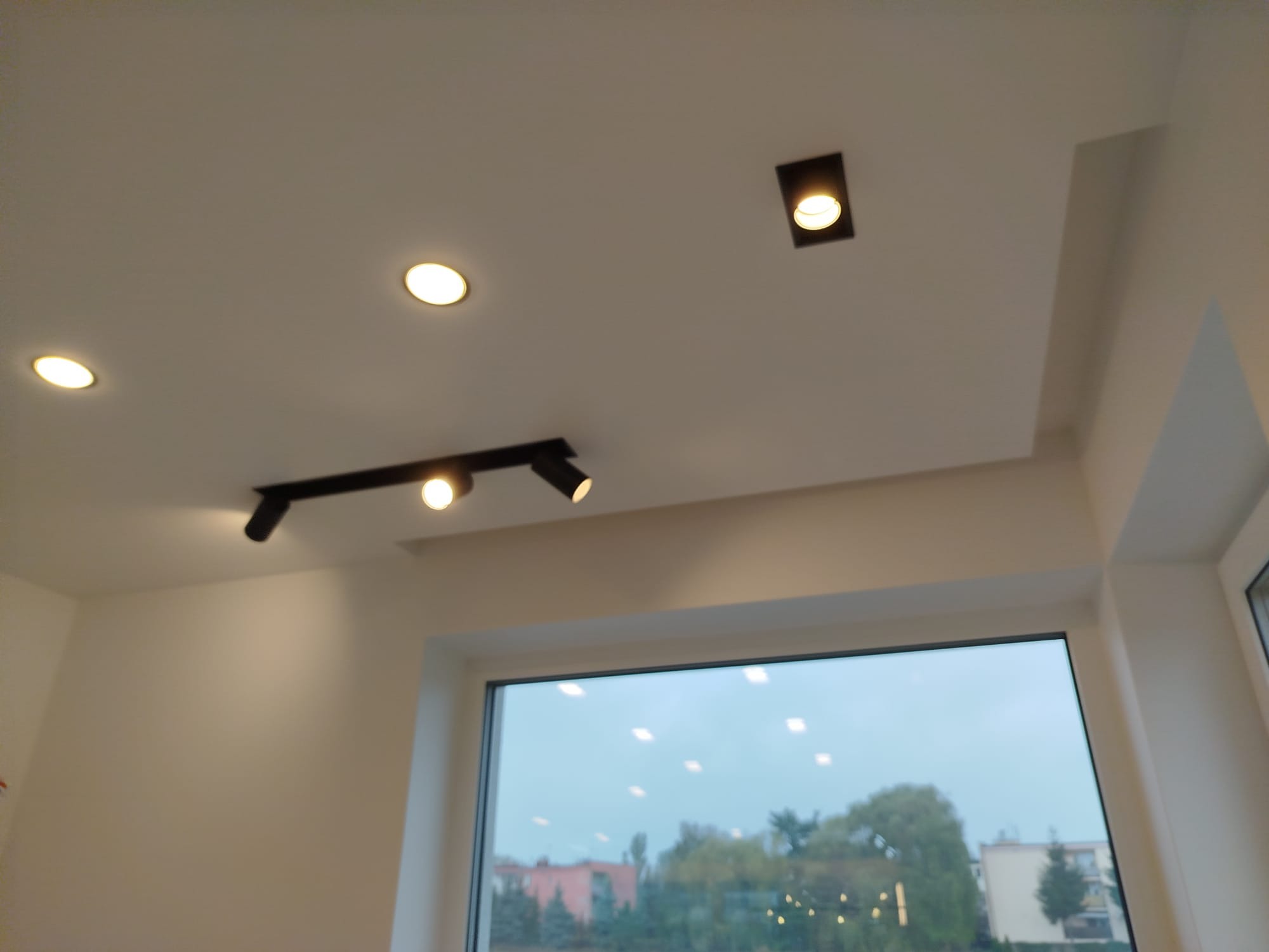 Biały sufit z zamontowanym oświetleniem punktowym: czarna szyna z reflektorami i okrągłe lampy wpuszczane. Widok na okno i budynki na zewnątrz.