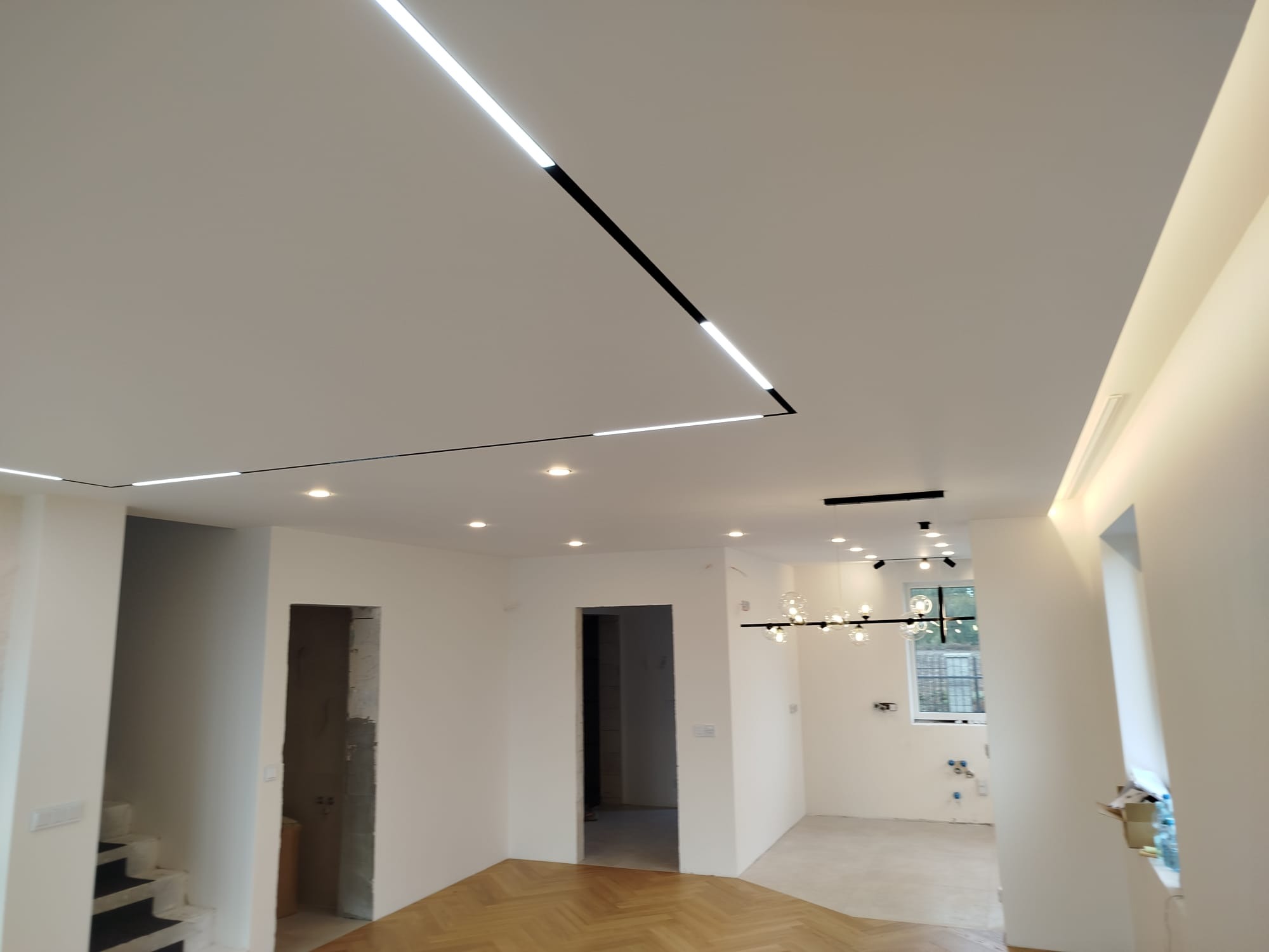 Białe wnętrze po remoncie z parkietem w jodełkę. Sufit podwieszany z nowoczesnym oświetleniem LED w geometrycznym układzie. Widoczne drzwi bez ościeżnic.