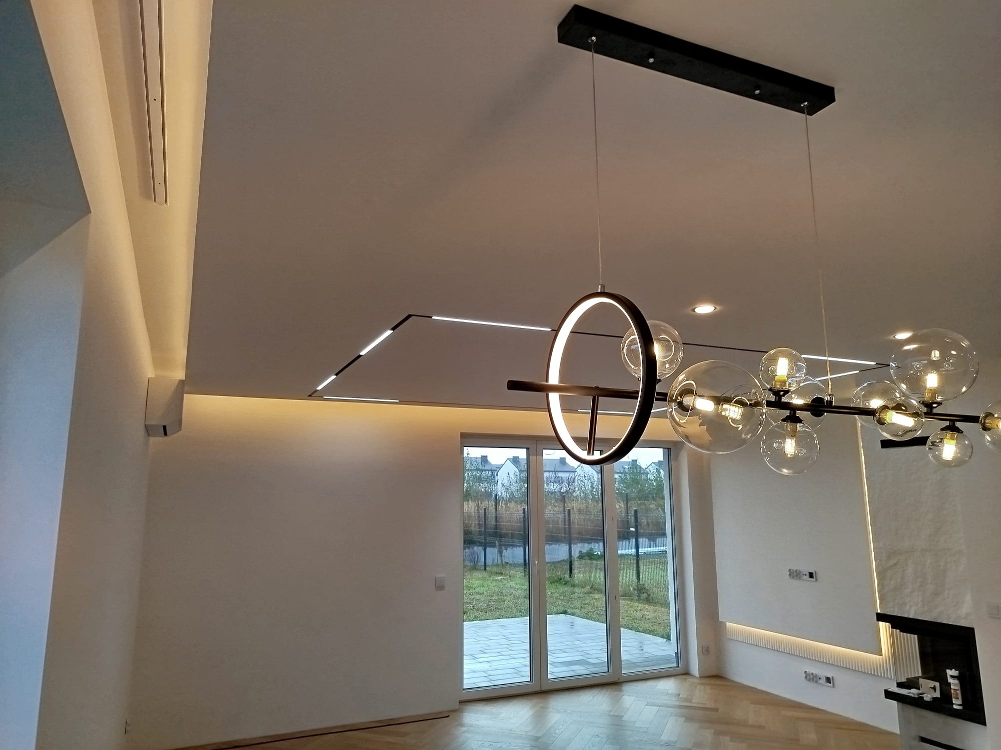 Nowoczesne oświetlenie sufitowe LED i designerska lampa wisząca w jasnym wnętrzu z parkietem i dużym oknem tarasowym. Minimalistyczny design.