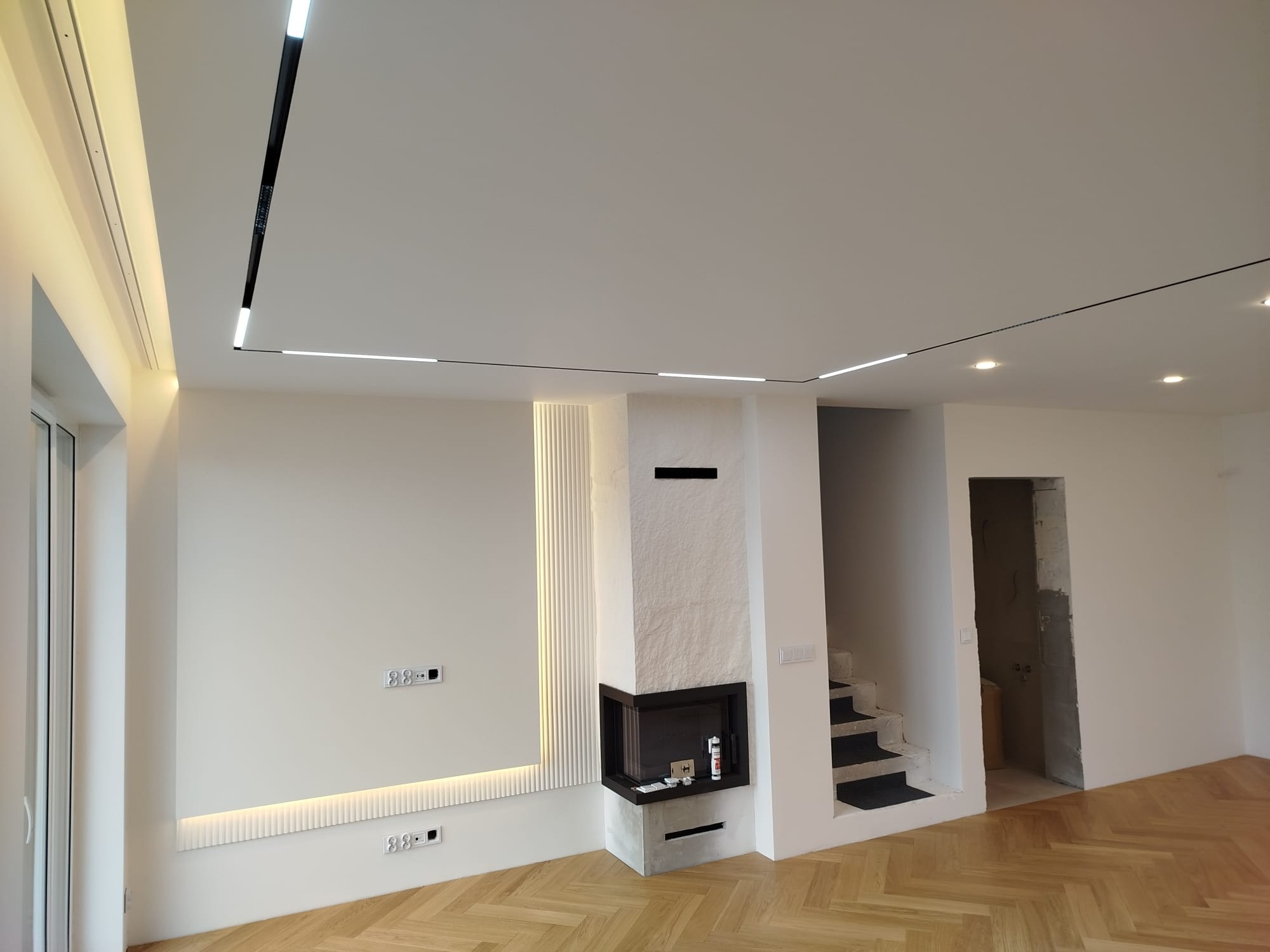 Nowoczesne wnętrze z parkietem w jodełkę, oświetleniem liniowym na suficie i kominkiem obłożonym kamieniem. Widoczne schody i minimalistyczne wykończenie ścian.