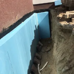 Niebieskie płyty izolacyjne XPS przy fundamentach budynku, częściowo zasypane ziemią, z widocznymi cegłami brukowymi na krawędzi wykopu i tekstem RTXPS300SLF 100x600x1250 na powierzchni płyt.