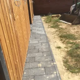 Ścieżka z szarych kostek brukowych ułożona wzdłuż drewnianej ściany budynku, z widoczną koparką w tle na piaszczystym terenie.