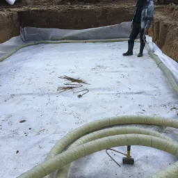 Wykop pod fundamenty wyłożony geowłókniną i drenażem, pracownik w kaloszach stoi na dnie wykopu, w tle żółta koparka i zabudowa jednorodzinna.