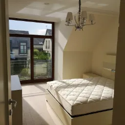 Jasna sypialnia na poddaszu z beżowymi panelami podłogowymi, łóżkiem z kremowym materacem i widokiem na osiedle domów przez duże okno balkonowe.