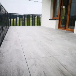 Jasnoszare płytki imitujące drewno na balkonie w Wiśniowej, z widoczną czarną balustradą i częściowo otwartymi drewnianymi drzwiami balkonowymi.