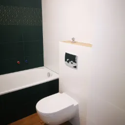 Nowoczesna łazienka z podwieszaną toaletą, ciemnozielonymi płytkami z geometrycznym wzorem i drewnopodobną podłogą, widok na wannę z armaturą.