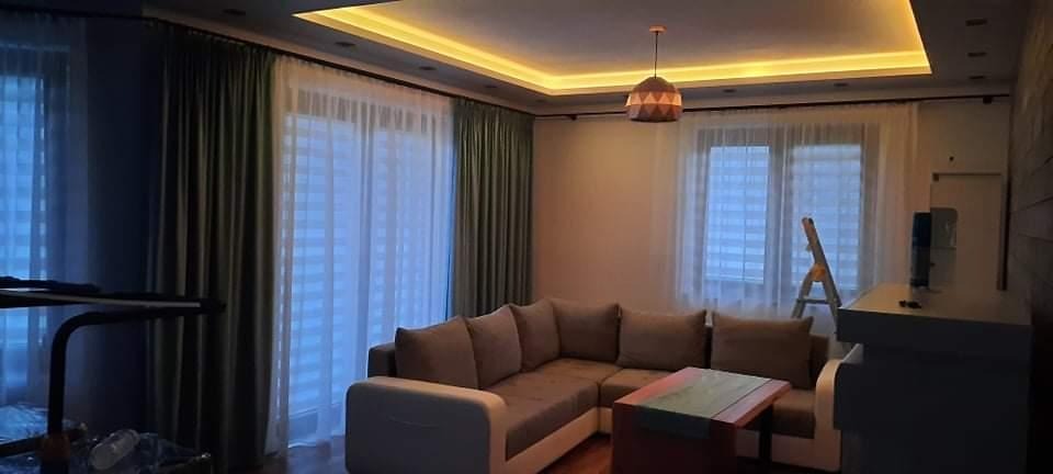 Salon w Wiśniowej z szarym narożnikiem, jasnymi zasłonami i podświetlanym sufitem, widok na okno z roletami.