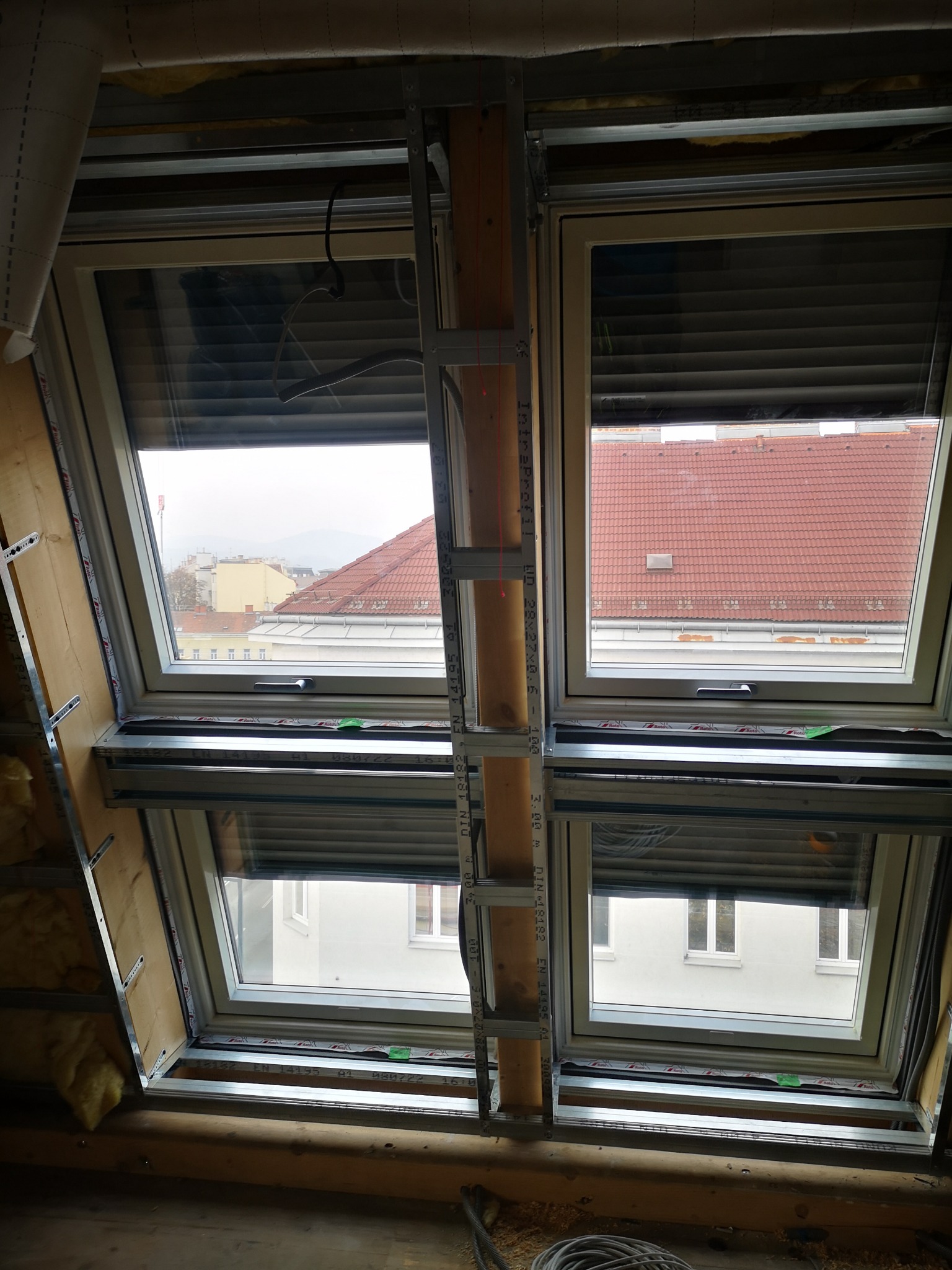 Dwa okna dachowe zamontowane w skośnym dachu, widok od wewnątrz podczas montażu z widocznymi elementami konstrukcyjnymi i częściowo zamontowanymi roletami zewnętrznymi. Za oknami widoczny budynek...
