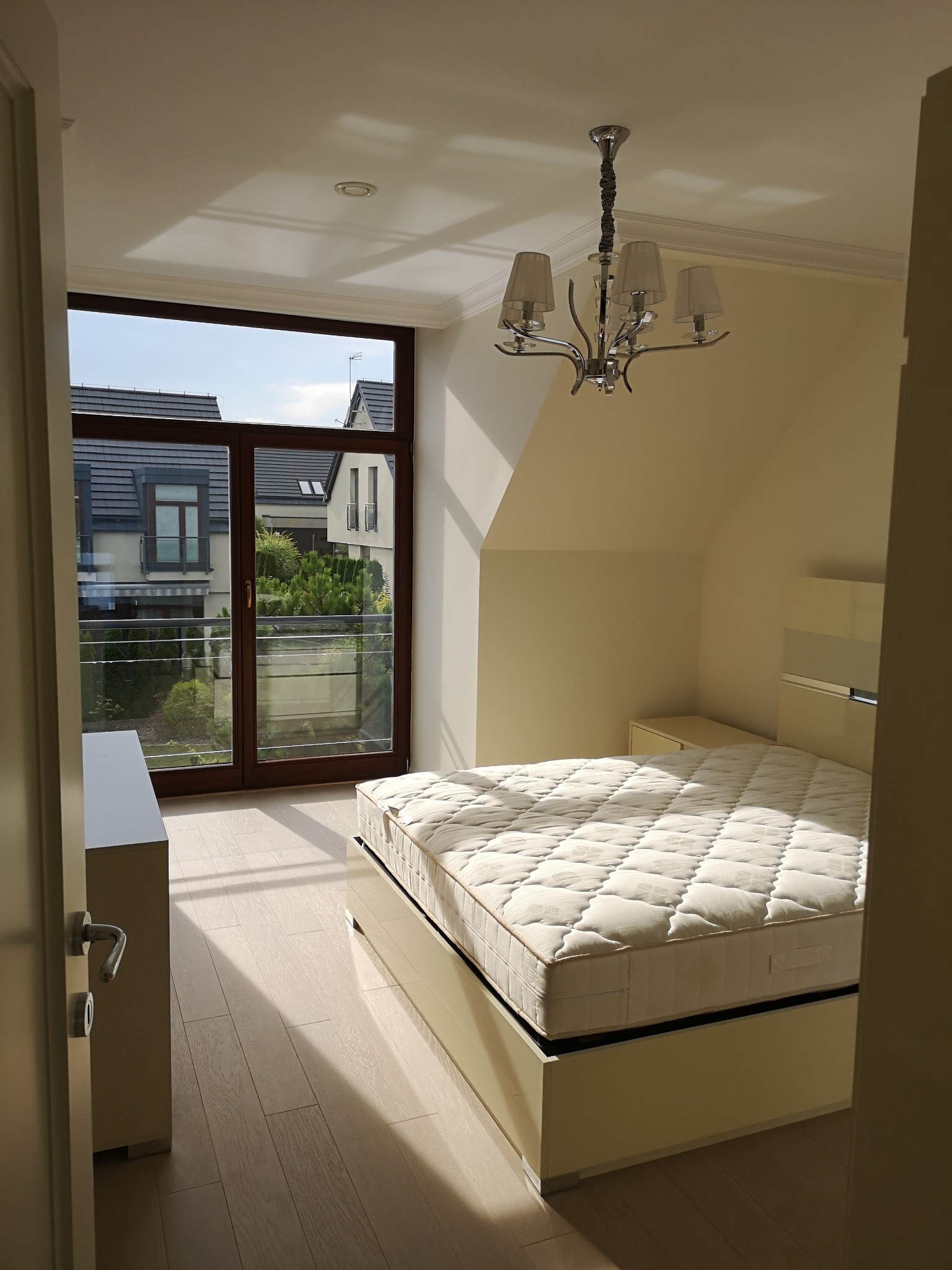 Jasna sypialnia na poddaszu z beżowymi panelami podłogowymi, łóżkiem z kremowym materacem i widokiem na osiedle domów przez duże okno balkonowe.