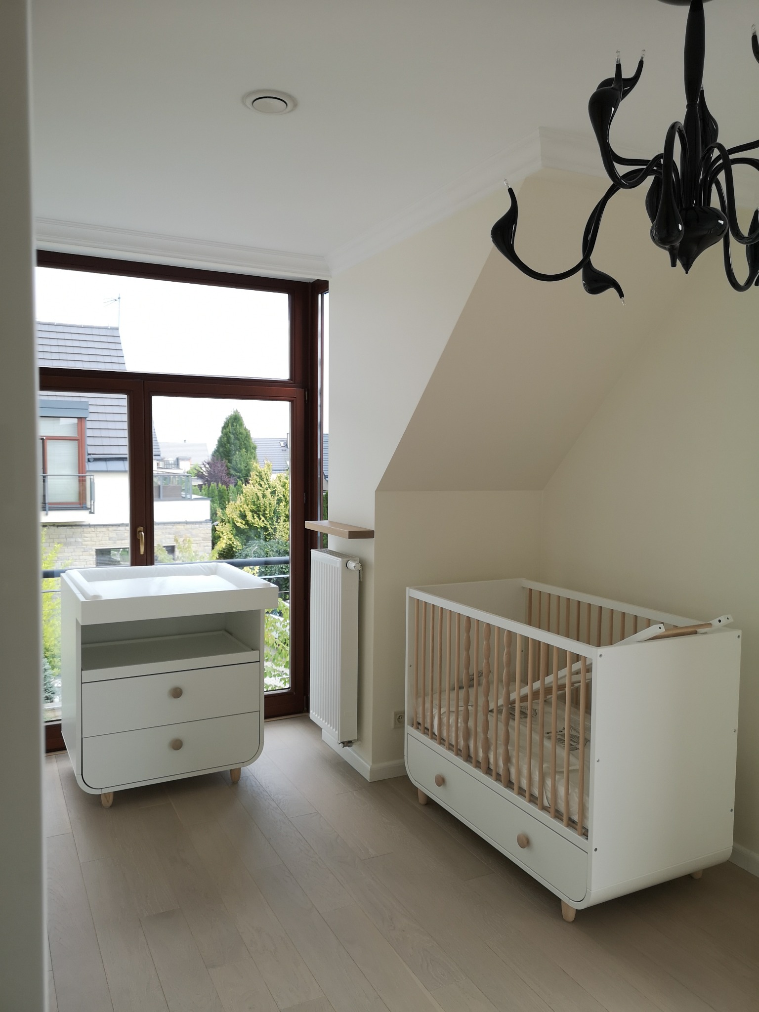 Jasne wnętrze pokoju dziecięcego na poddaszu z drewnianą podłogą, białym łóżeczkiem ze szczebelkami i komodą z przewijakiem, doświetlone dużym oknem z widokiem na zieleń.