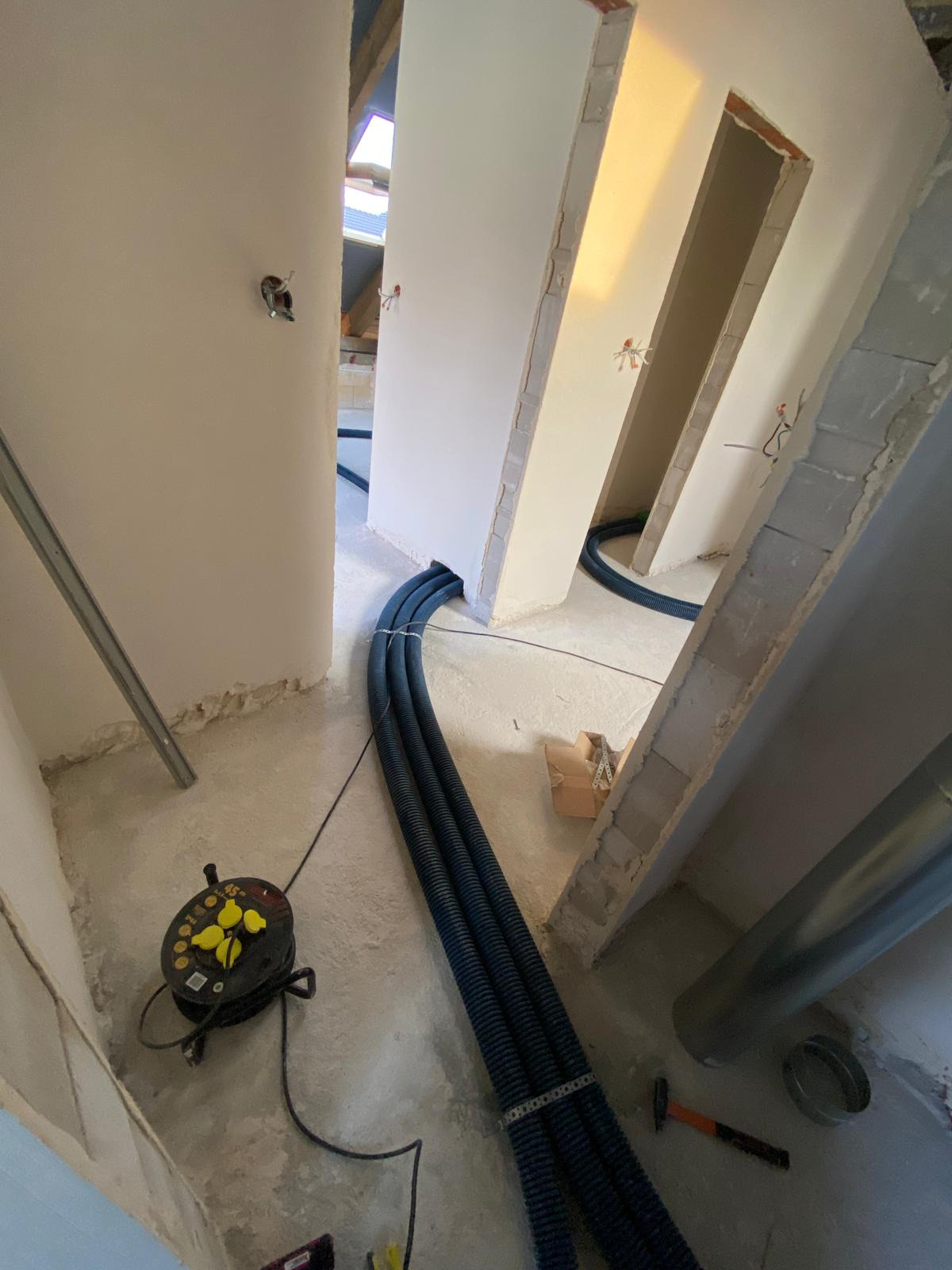 Wnętrze w trakcie prac wykończeniowych, widoczne trzy czarne, karbowane rury wentylacyjne prowadzone wzdłuż ściany, zwinięty przedłużacz na podłodze, fragment poddasza z oknem dachowym w tle.