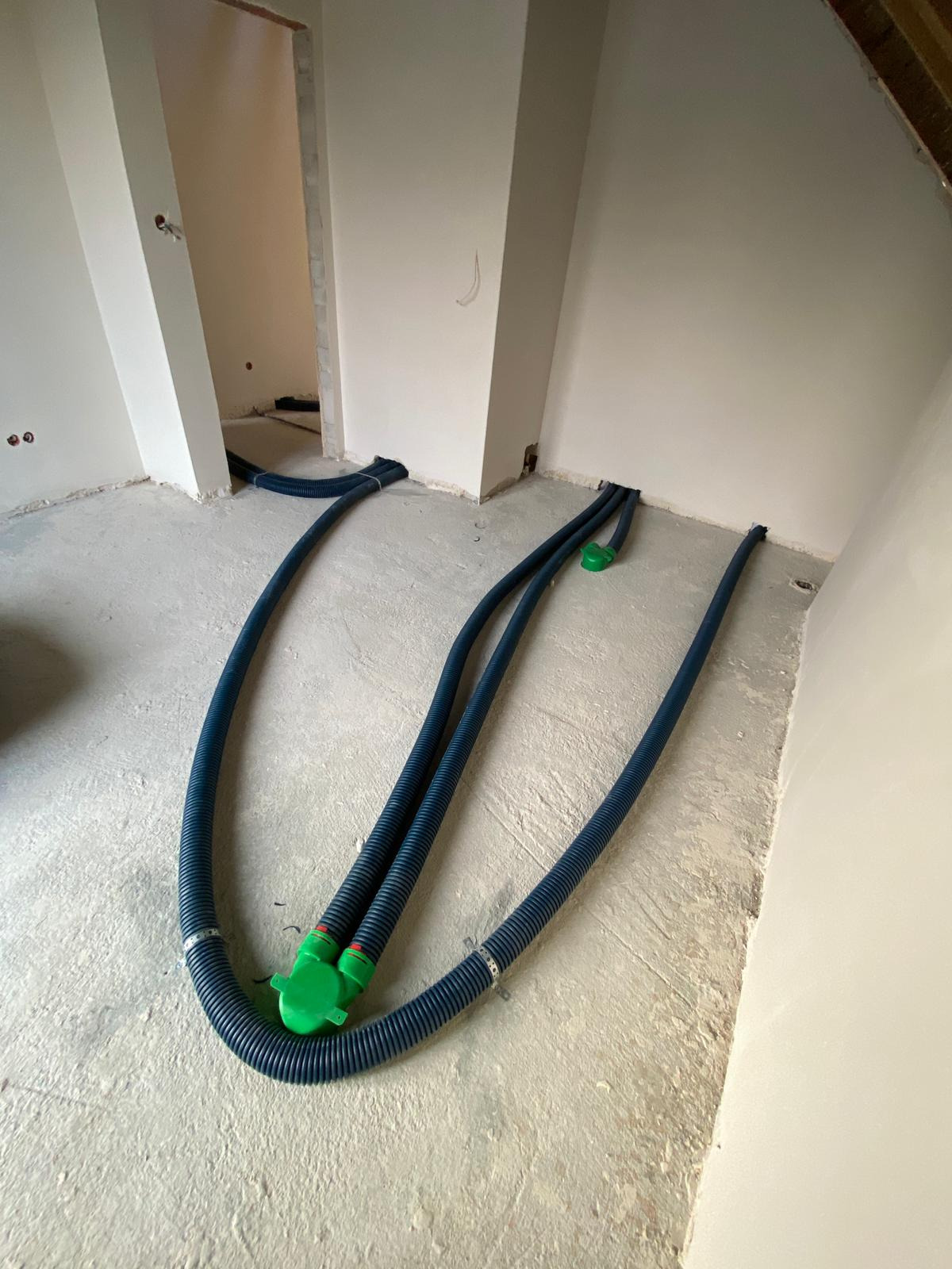 Niebieskie, karbowane rury wentylacyjne z zielonymi łącznikami na surowym, betonowym podłożu w trakcie instalacji rekuperacji w nowym budynku.