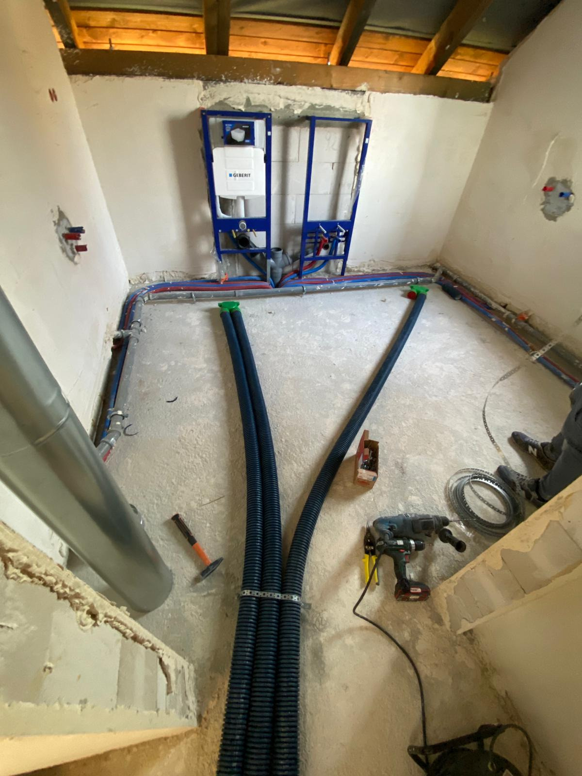 Surowy stan pomieszczenia w trakcie prac instalacyjnych: widoczne rury wentylacyjne w niebieskiej karbowanej osłonie, narzędzia (wiertarka, młotek), instalacja sanitarna Geberit na ścianie,...