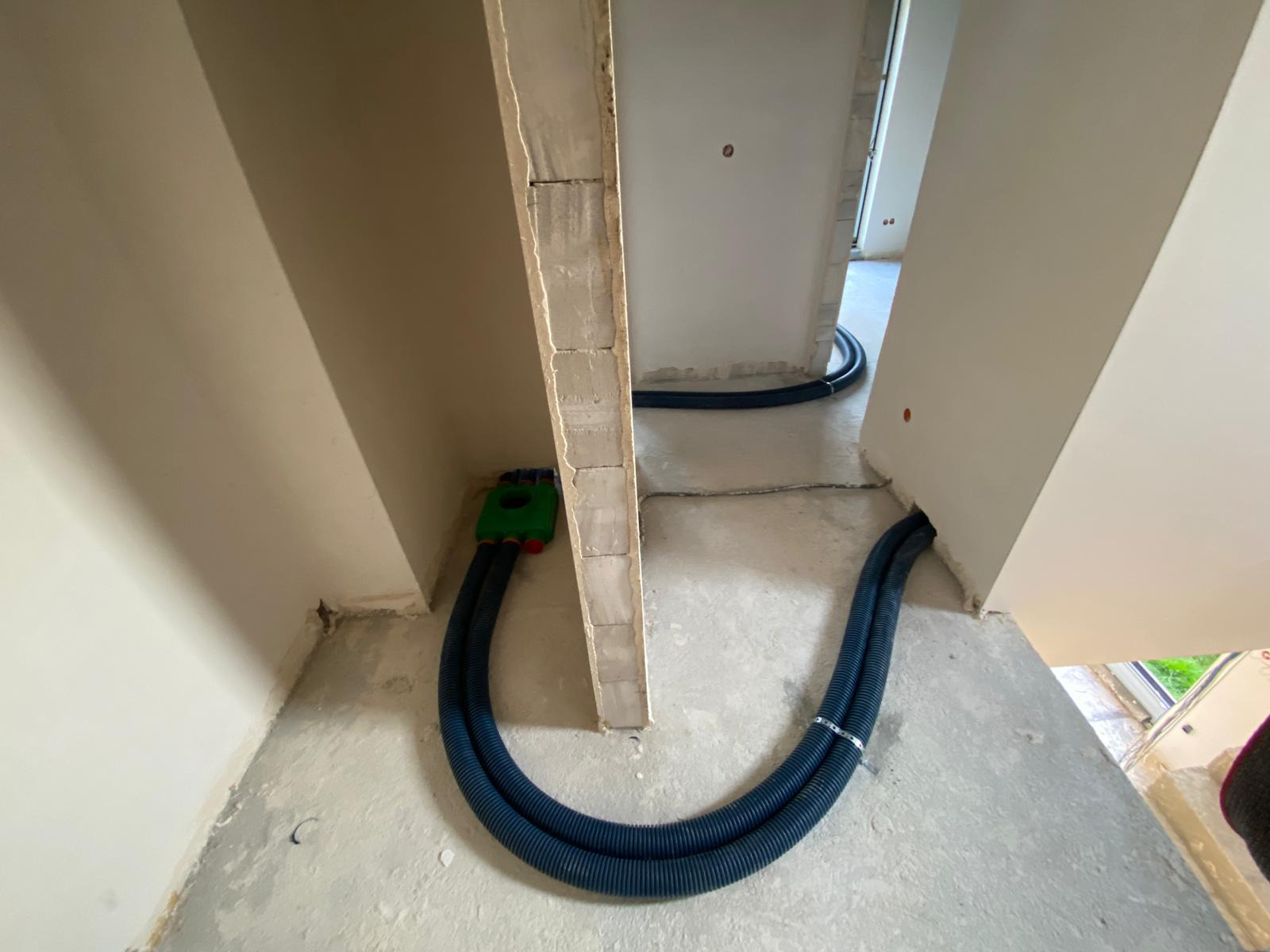 Dwa niebieskie, karbowane przewody wentylacyjne prowadzone wzdłuż surowej ściany z betonu komórkowego wewnątrz budynku w stanie deweloperskim, połączone z zieloną skrzynką rozdzielczą.