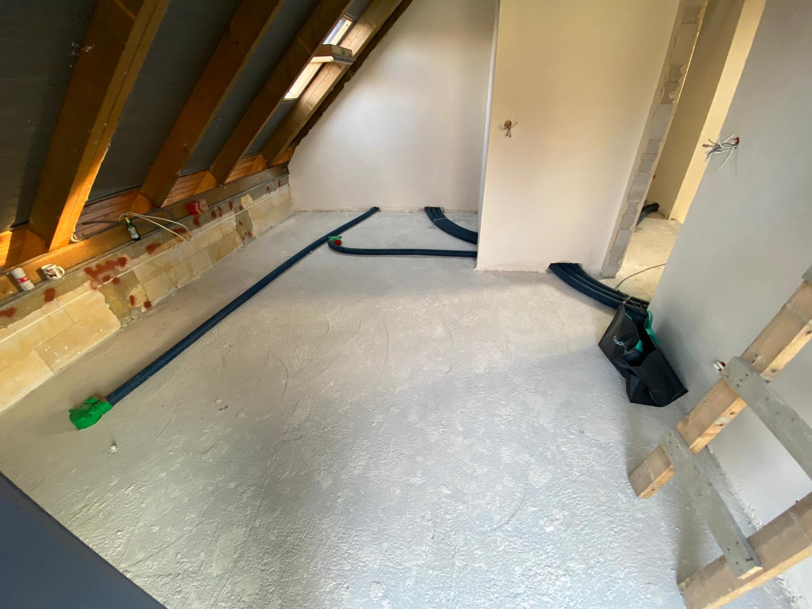 Poddasze w trakcie prac wykończeniowych, widoczne rury wentylacyjne w kolorze czarnym z zielonymi złączkami, ułożone na szarej wylewce, z odsłoniętą konstrukcją dachu i częściowo otynkowanymi...