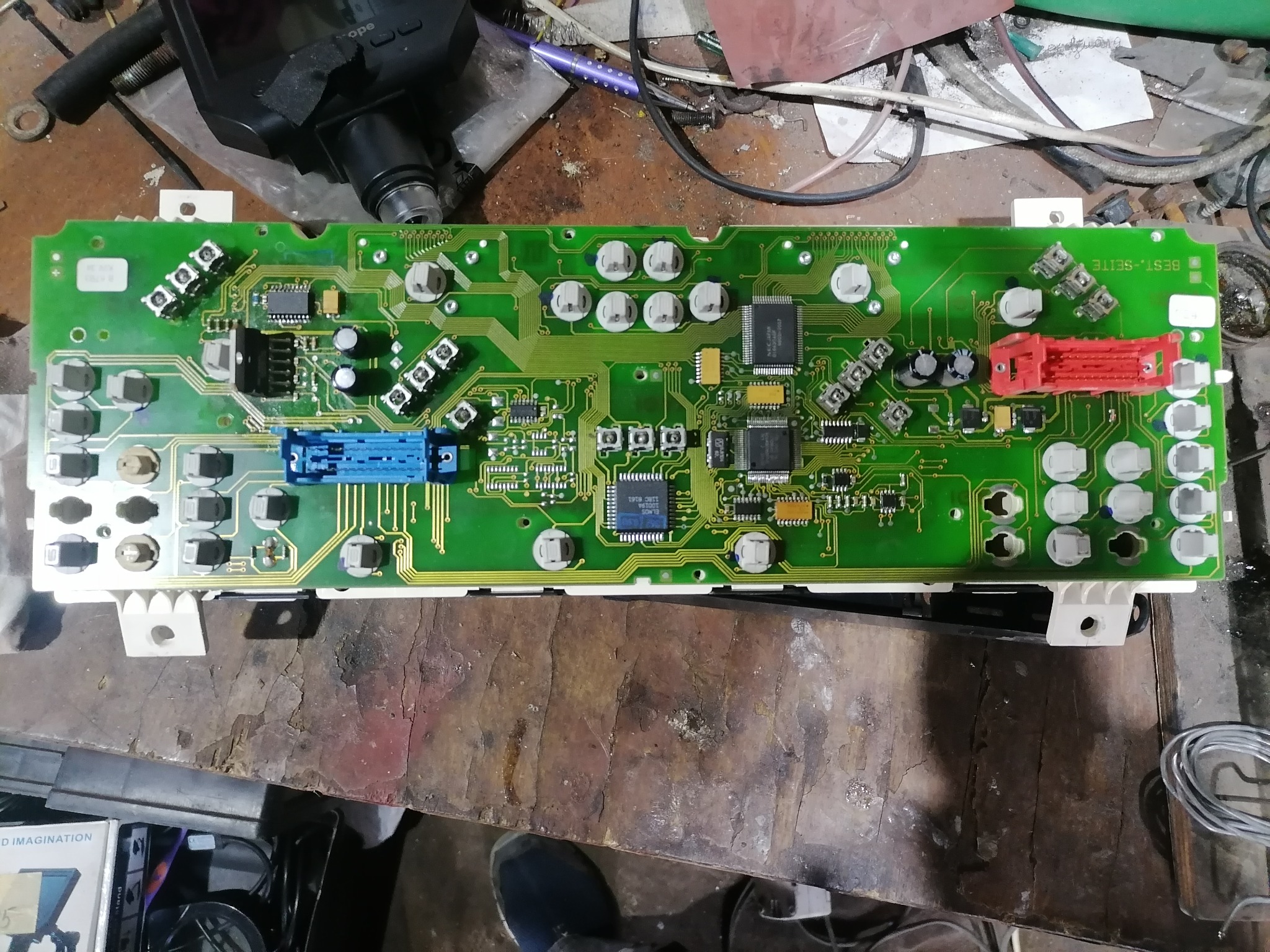 Zielona płytka drukowana deski rozdzielczej samochodu z elementami elektronicznymi, widoczna na warsztatowym stole, z przewodami i narzędziami w tle.