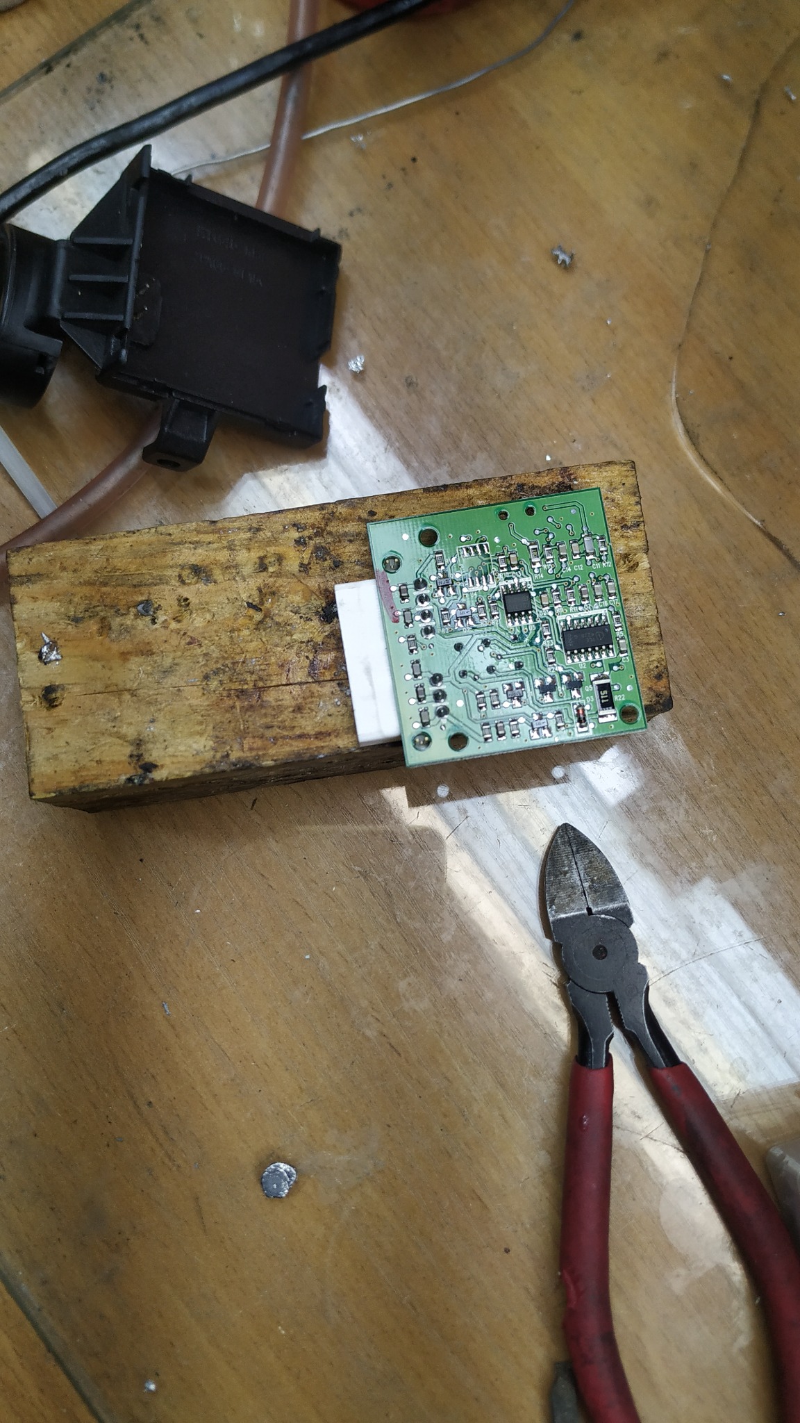 Zielona płytka drukowana z elementami elektronicznymi, umieszczona na drewnianym klocku, obok leżą obcęgi z czerwonymi rączkami i czarna obudowa elementu samochodowego, na drewnianym blacie.
