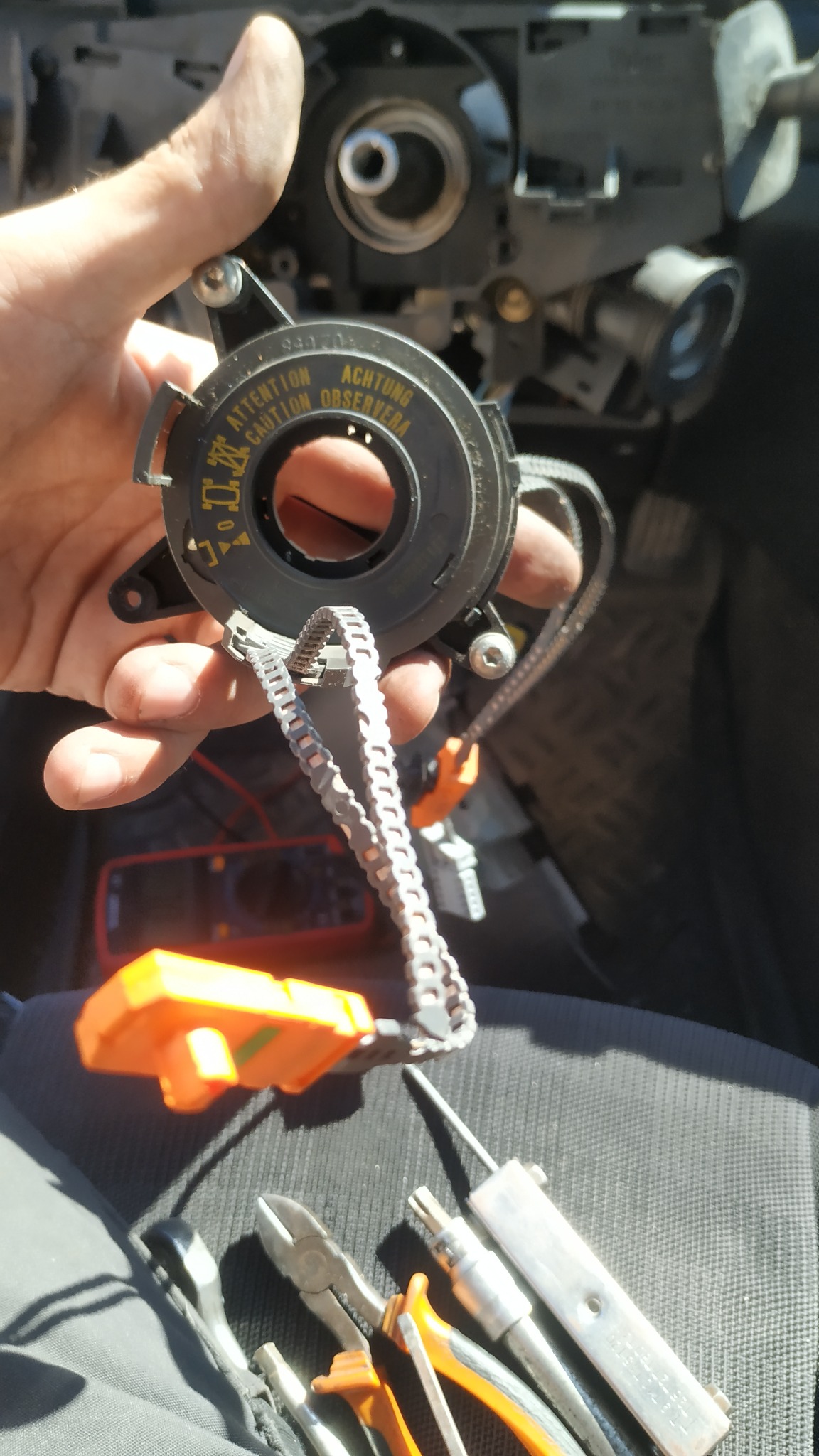 Demontaż modułu poduszki powietrznej kierownicy z ostrzeżeniem 'Attention Caution Observa' w samochodzie, widoczne narzędzia i wnętrze kokpitu.