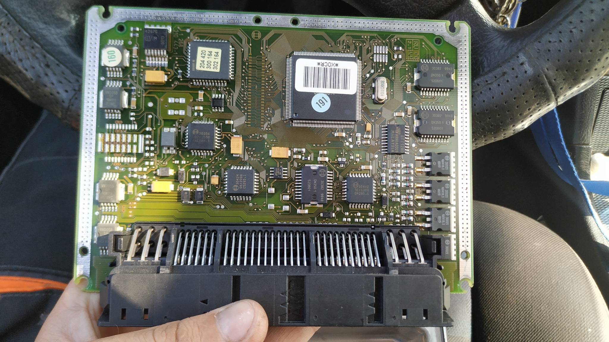 Otwarty sterownik silnika samochodowego z widocznymi elementami elektronicznymi, układami scalonymi i pinami, trzymany w dłoni na tle wnętrza pojazdu.