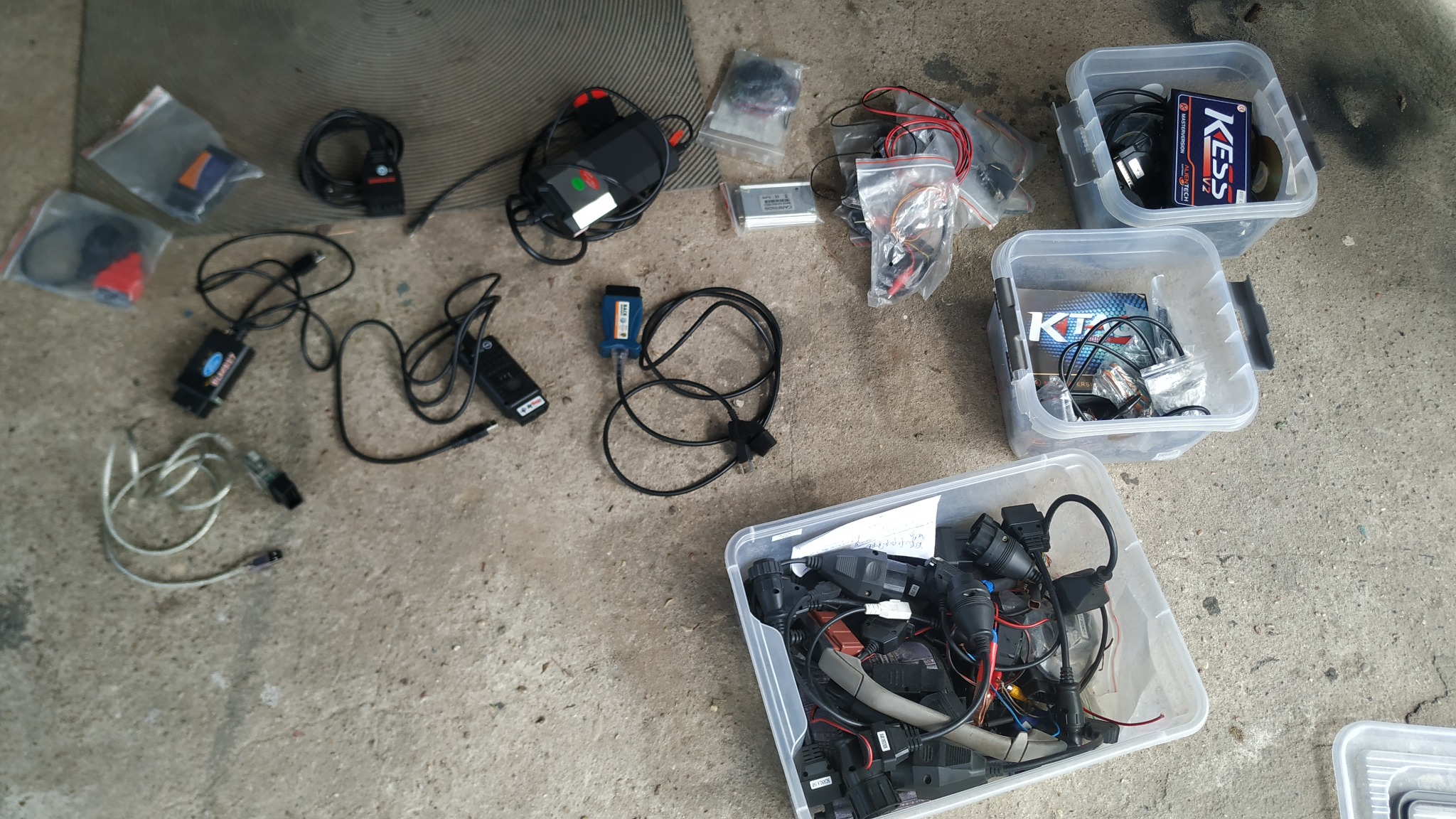 Rozłożone na betonowej posadzce narzędzia i akcesoria diagnostyczne do samochodów, w tym interfejsy diagnostyczne, kable i adaptery, niektóre z nich w przezroczystych plastikowych pojemnikach...