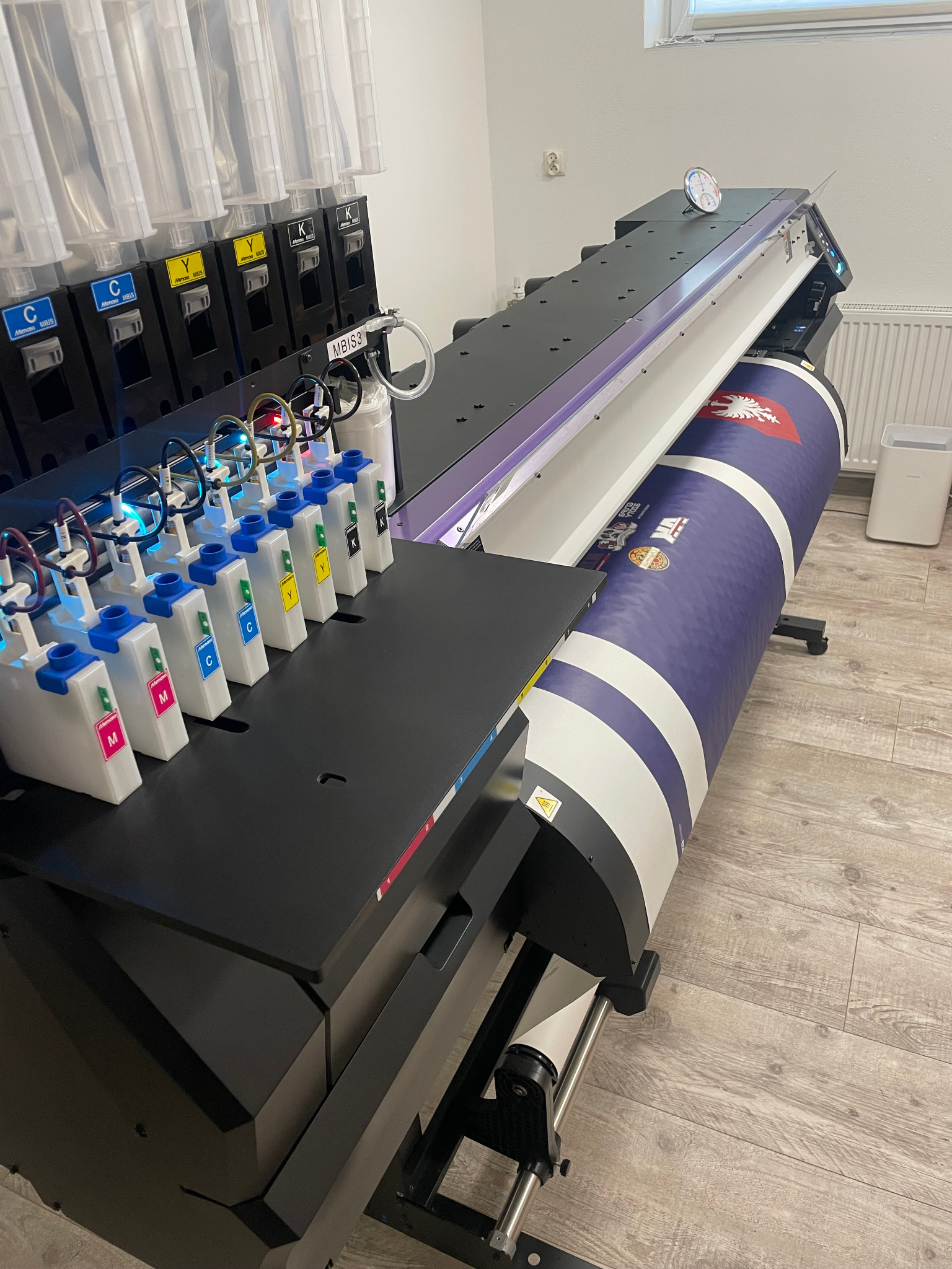 Szerokokątne ujęcie drukarki wielkoformatowej z pełnymi pojemnikami na tusze CMYK, drukującej baner z herbem i logotypami na podłodze z paneli.