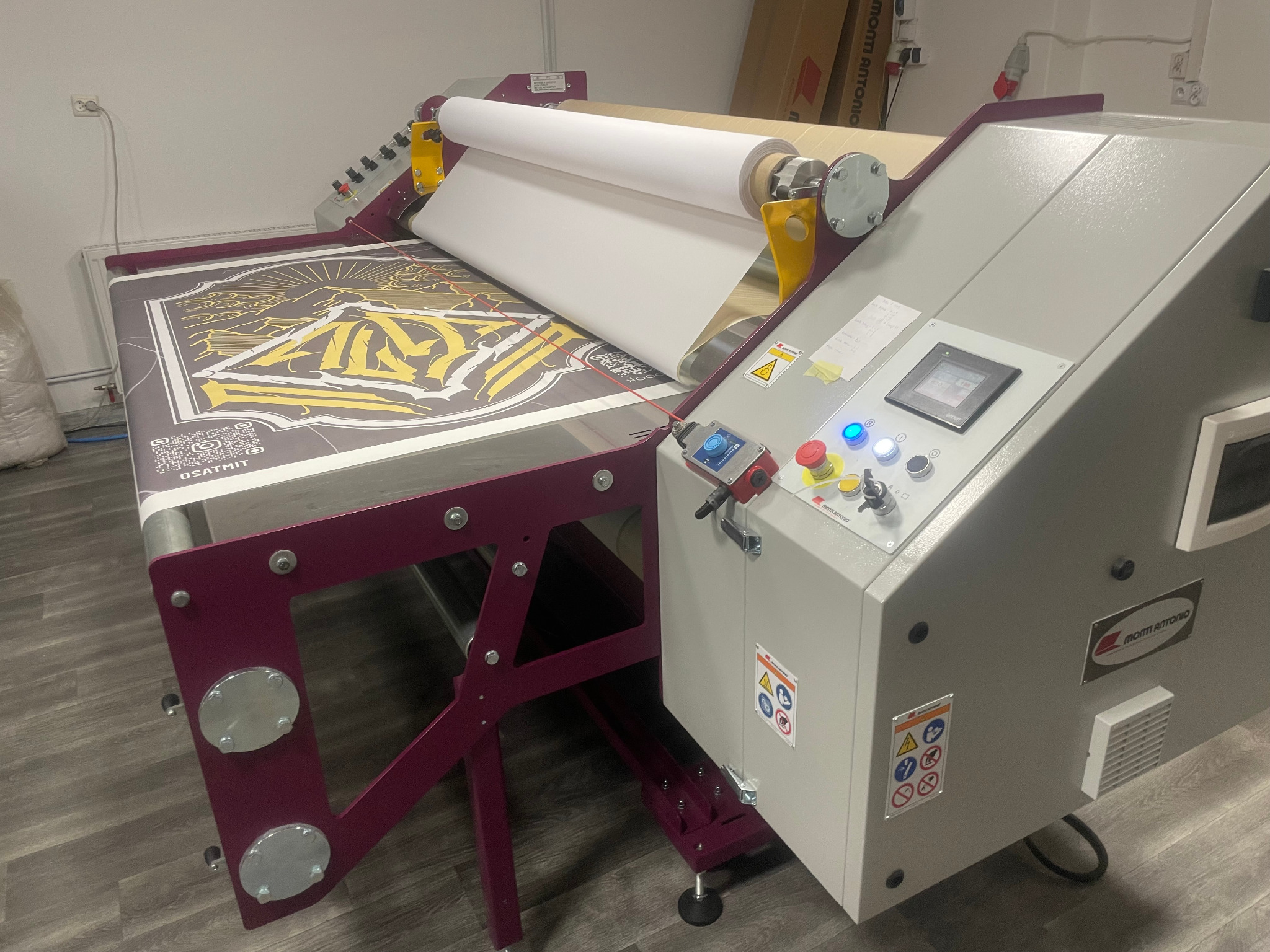 Przemysłowa maszyna do nadruku na tkaninach z rolką białego materiału, częściowo zadrukowanym panelem z motywem graficznym w odcieniach szarości i żółci oraz panelem sterowania z przyciskami...